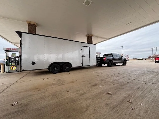 A black pickup truck is parked next to a large white enclosed trailer at a gas station, with a fueling pump nearby.