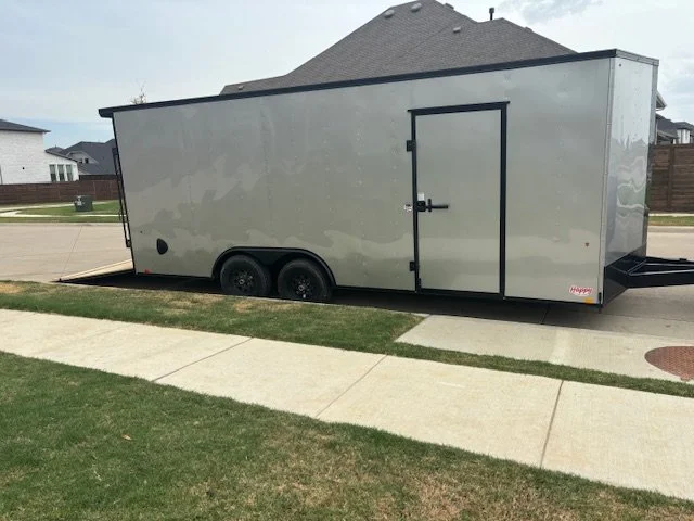 Gray enclosed trailer parked on suburban street with grass and sidewalk in front, residential houses in background.