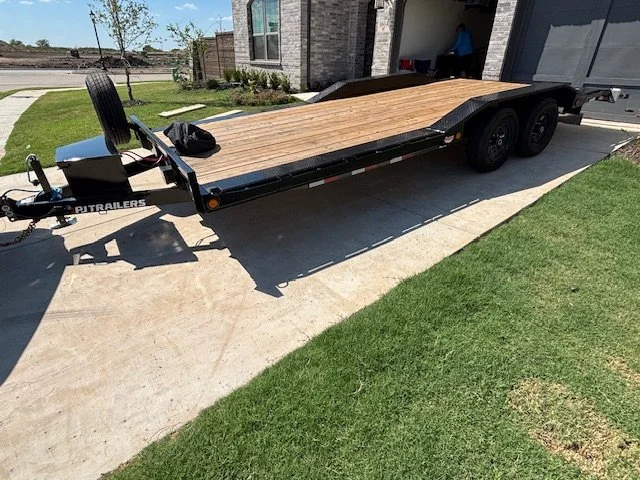 A flatbed trailer parked on a driveway next to a house, with a tire and some tools on the trailer.