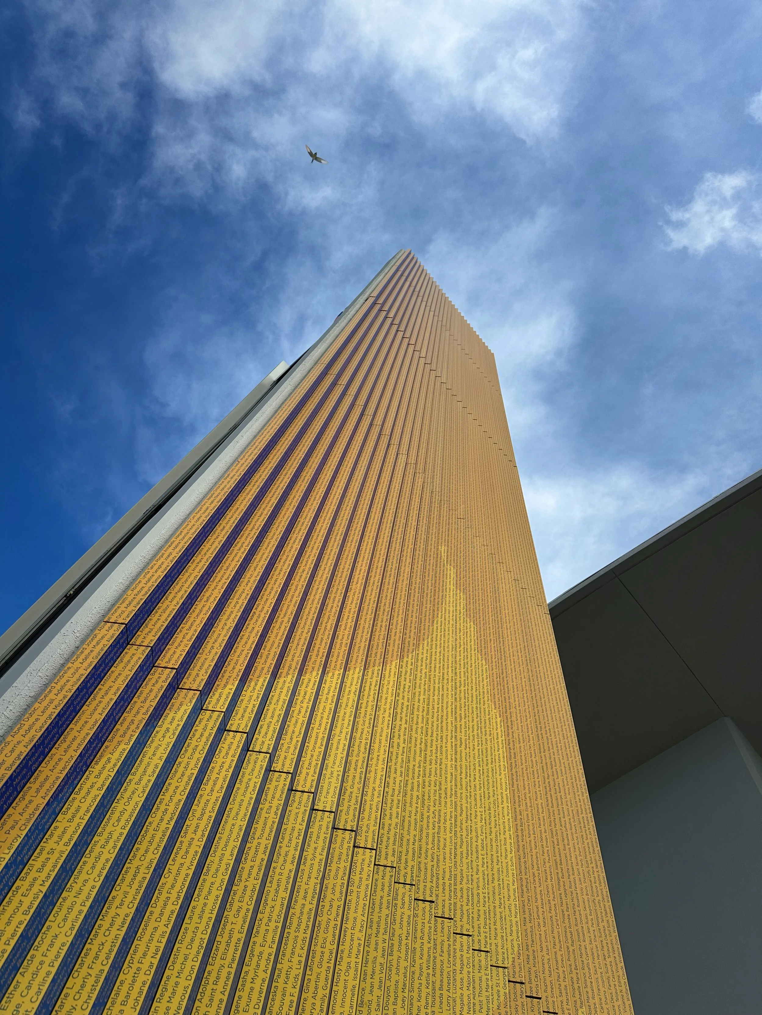 Looking up at a tall architectural feature element with a gold exterior and vertical lines, against a blue sky with scattered clouds and a bird flying overhead.