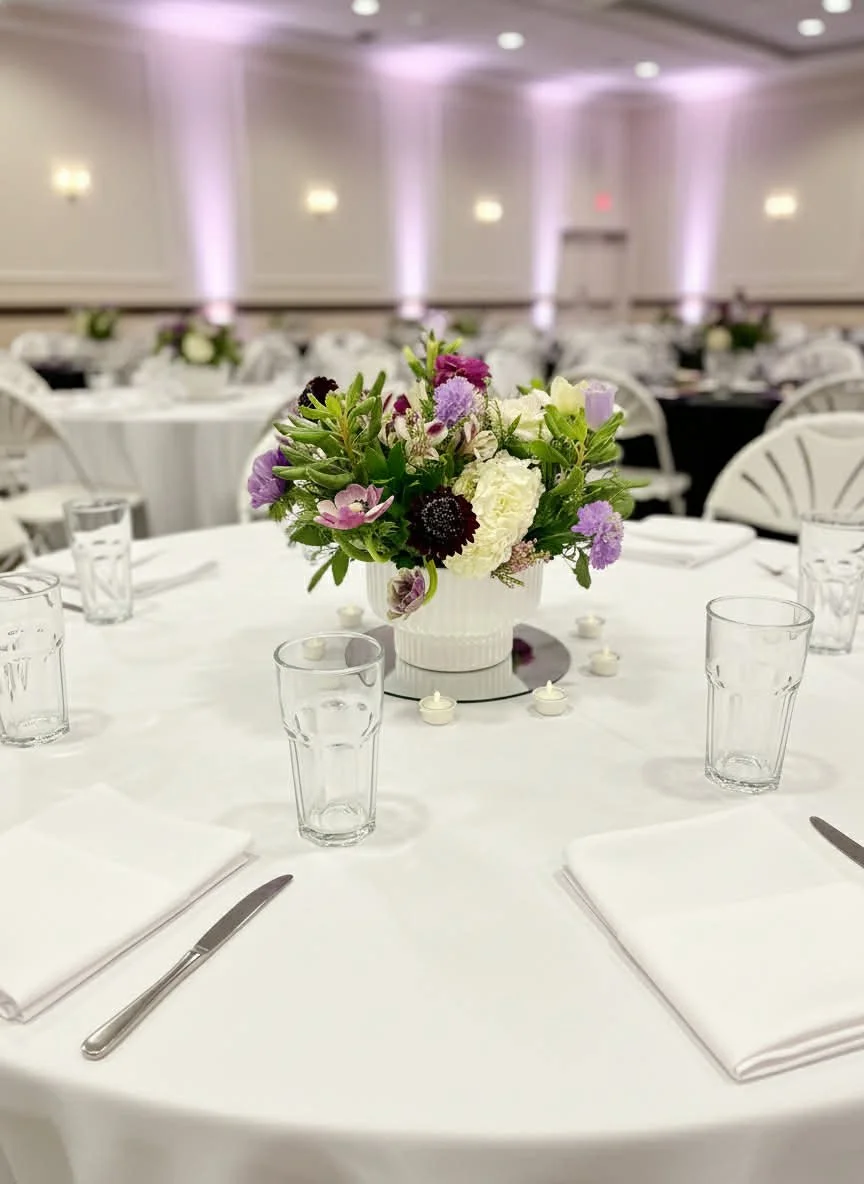 Wedding reception table with white tablecloth, floral centerpiece with purple, white, and dark flowers, surrounded by empty glasses, white napkins, and silverware, in a decorated banquet hall with pink uplighting.