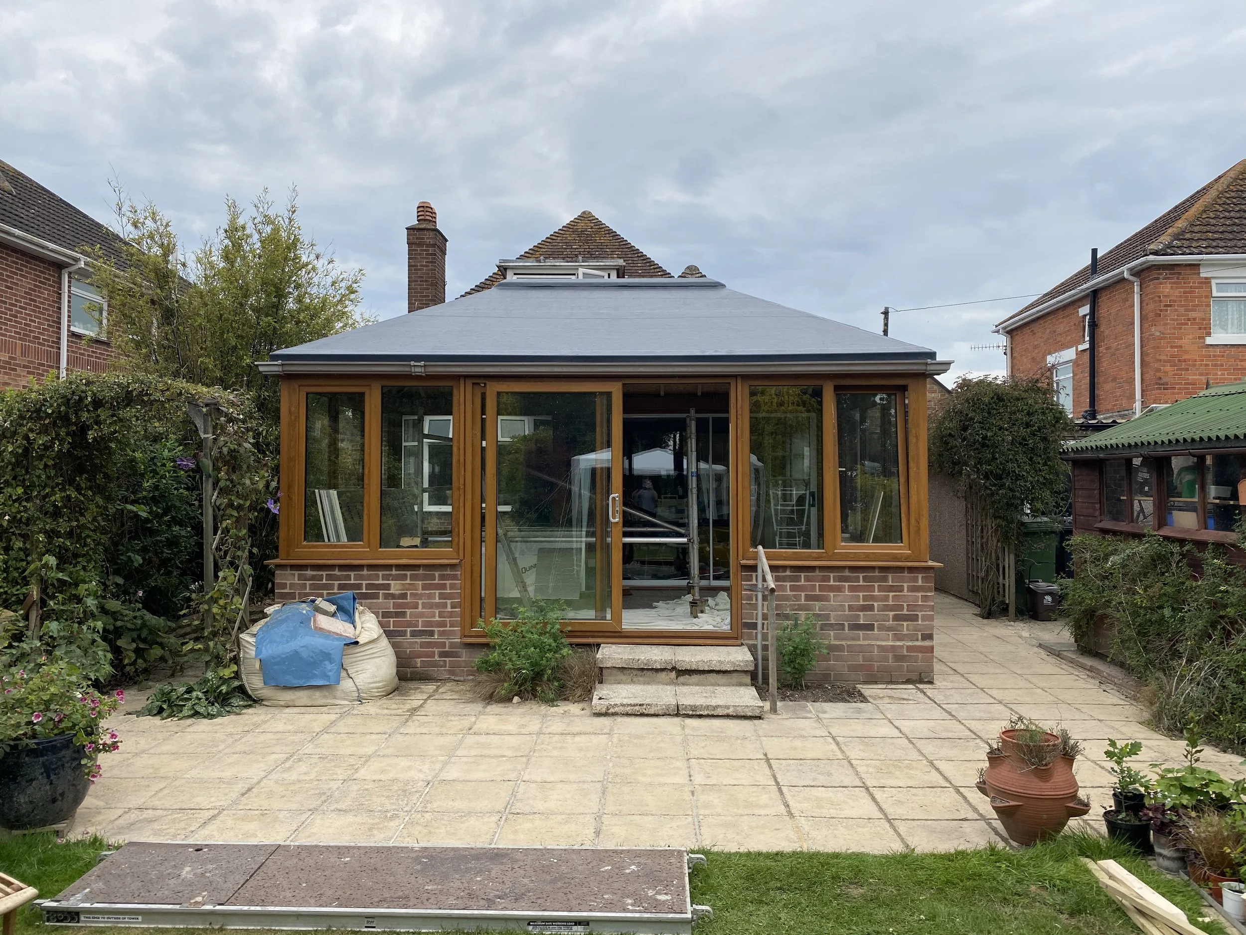 Backyard with a brick-built house featuring a glass conservatory, with potted plants, a bag, and construction materials in the yard.