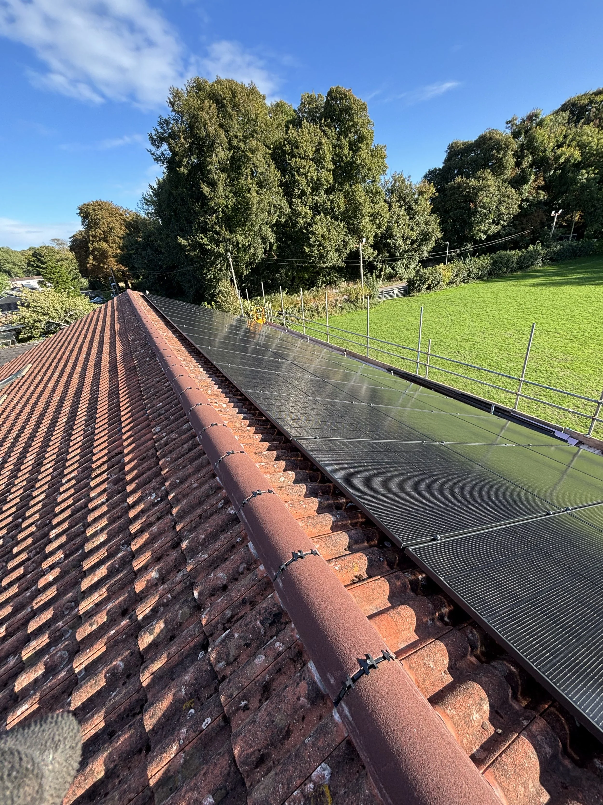 Solar panels installed on a red-tiled roof, with a green field and trees in the background under a blue sky.
