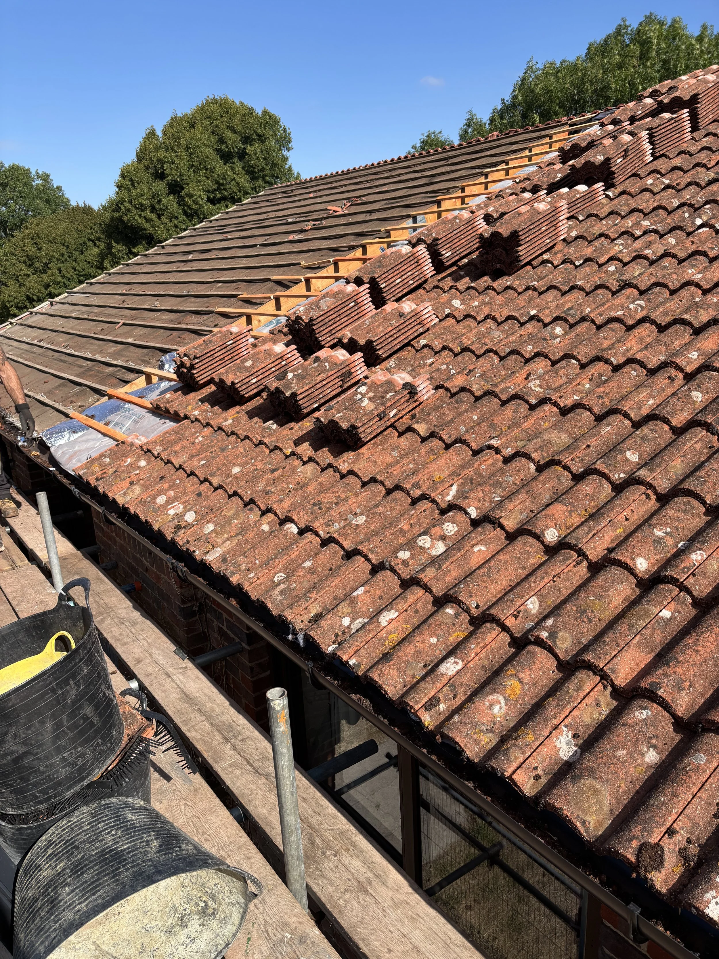 Roof with partially replaced red clay tiles, scaffolding on the side, and trees in the background.