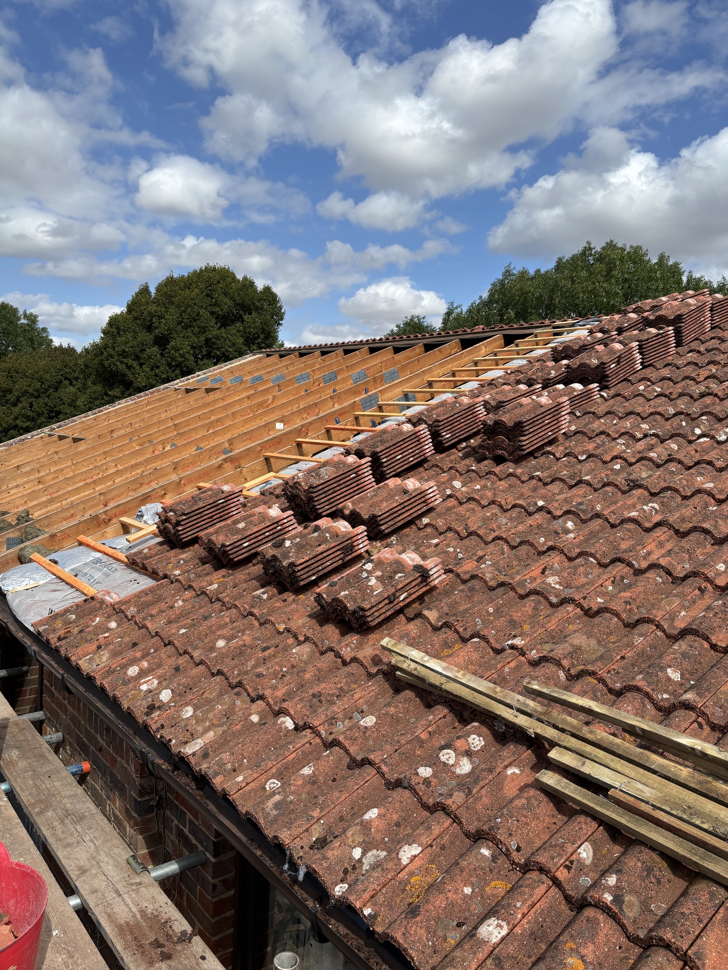 Front view of a residential roof under renovation with part of the original red brick tiles and the new wooden framework for new tiles, under a partly cloudy blue sky.