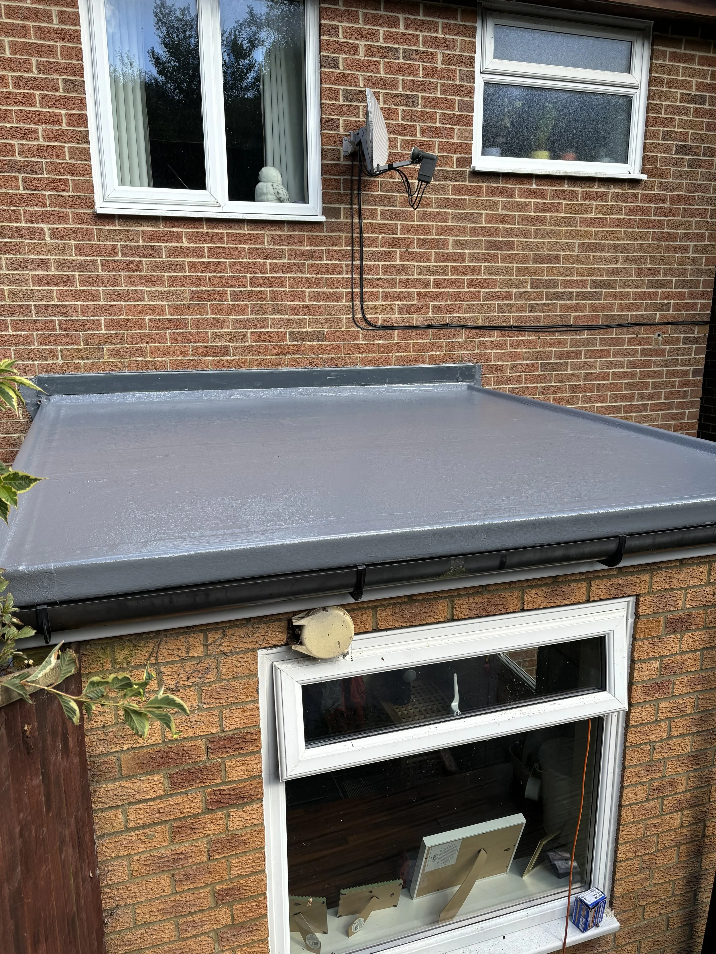 Exterior view of a brick house with two windows, one upstairs and one downstairs, and a gray roofed awning covering a window. A satellite dish is mounted on the side of the house. Some plants and a wooden fence are visible on the side.