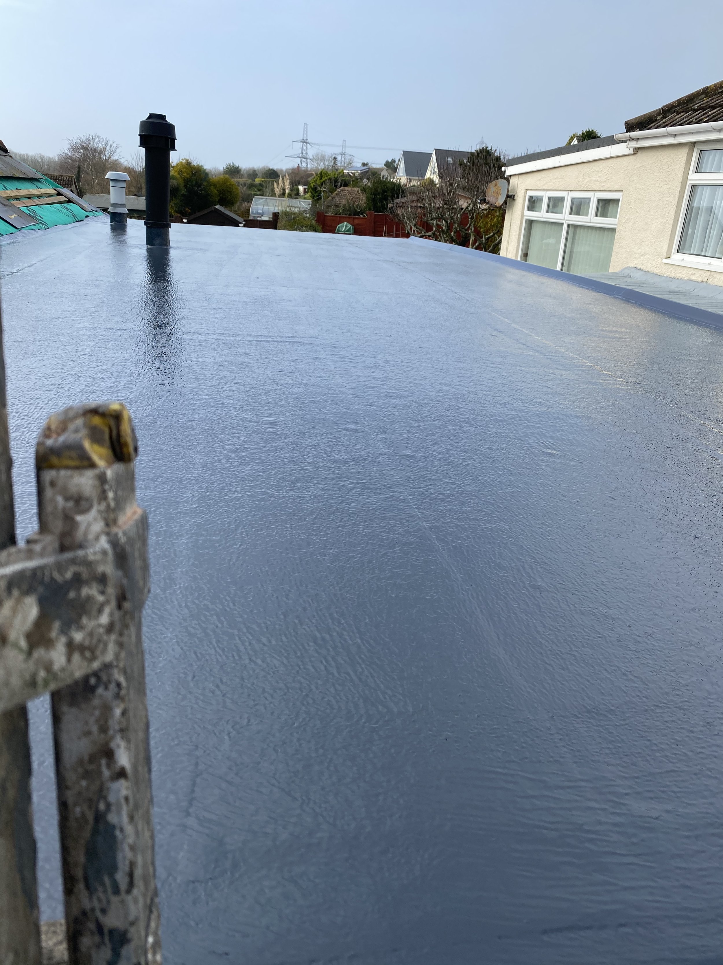 Flat roof covered with a dark waterproof coating with three vents and a chimney pipe, residential neighborhood with trees, houses, and sky