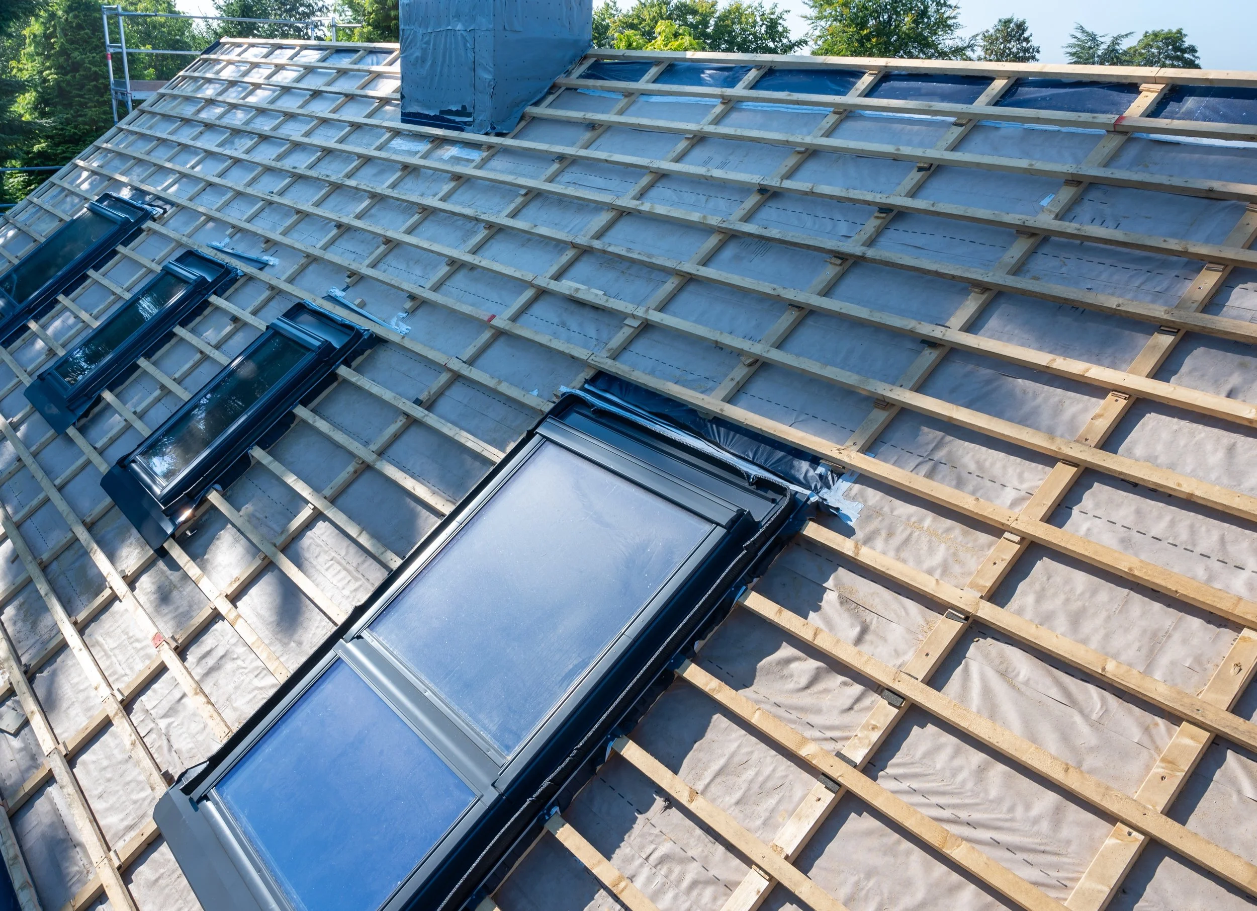 Roof under construction with wooden framing and installed skylights, showing a sloped roof with trees in the background.