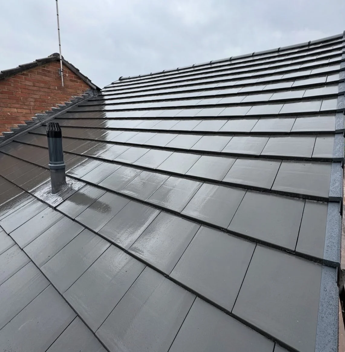 Newly installed dark gray roof tiles on a residential house with a chimney pipe and a brick wall in the background.
