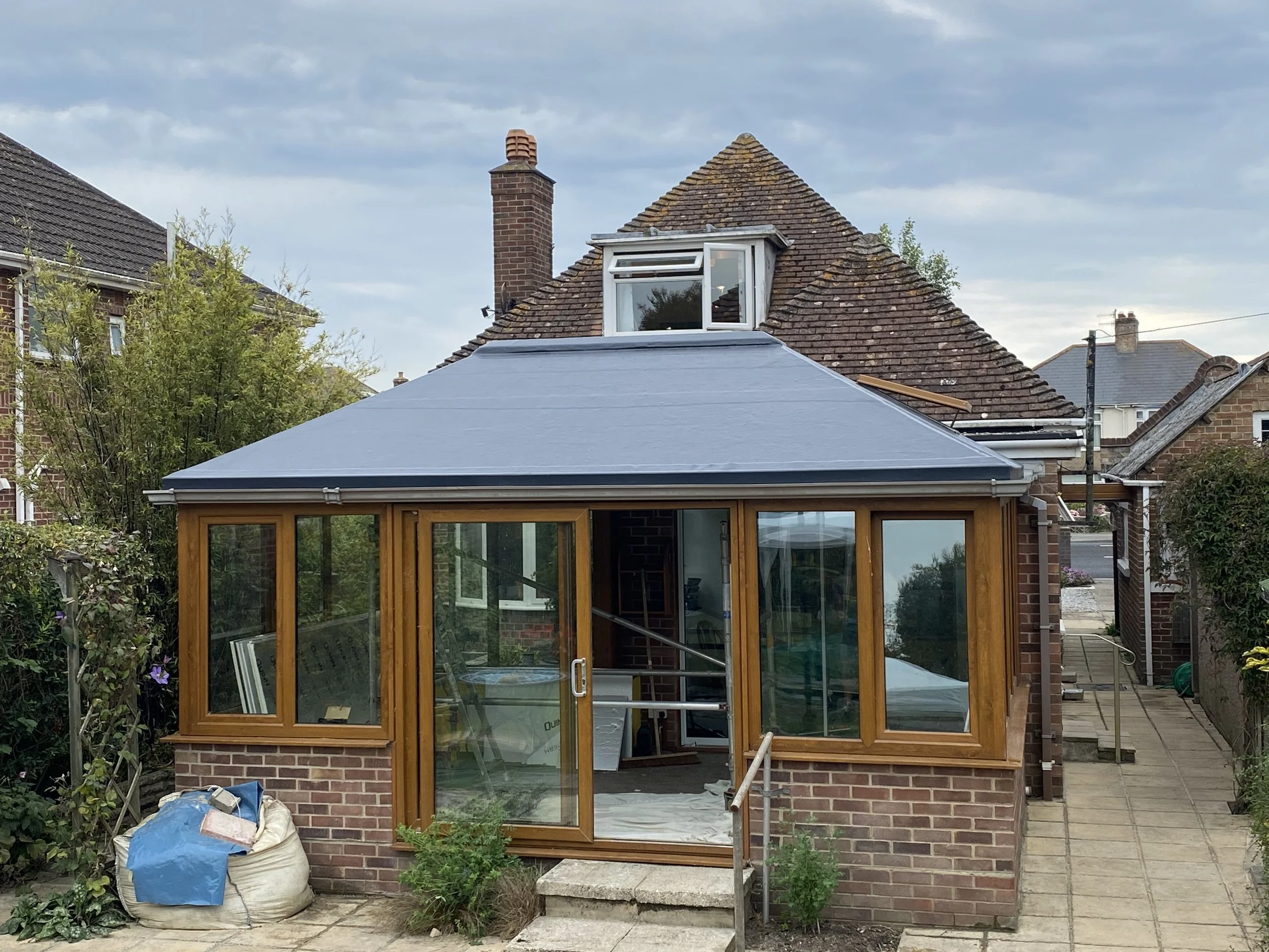 A small brick house with a newly constructed wooden and glass extension. The extension has a blue roof and sliding glass doors. There are construction materials and tools nearby, indicating ongoing work.