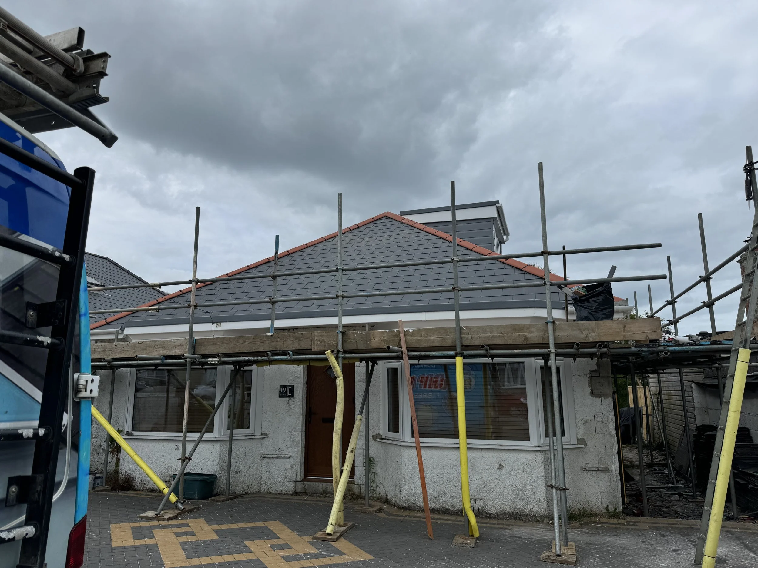House under renovation with scaffolding around roof, cloudy sky overhead, and construction equipment nearby.