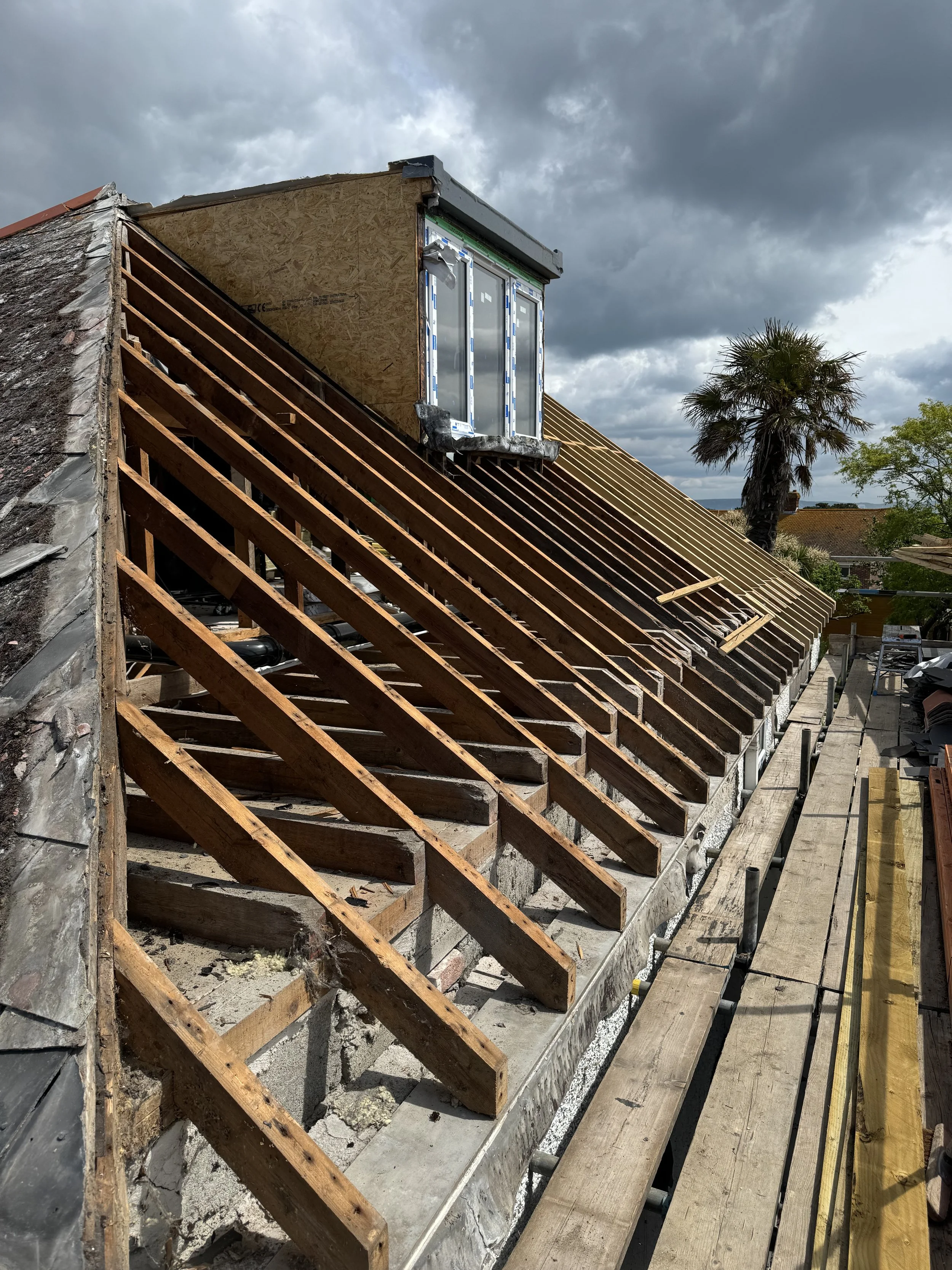 Roof under construction with exposed wooden rafters, a small window opening, and cloudy sky.