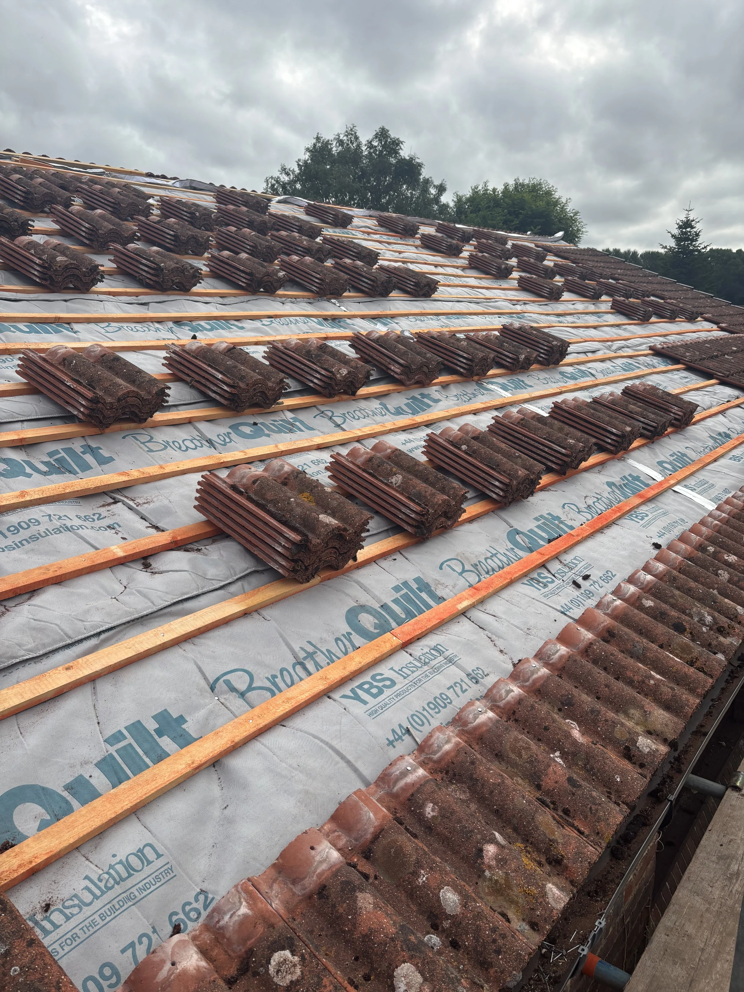 Roof under construction with wooden battens, roofing tiles, and underlayment material visible, with trees and cloudy sky in the background.