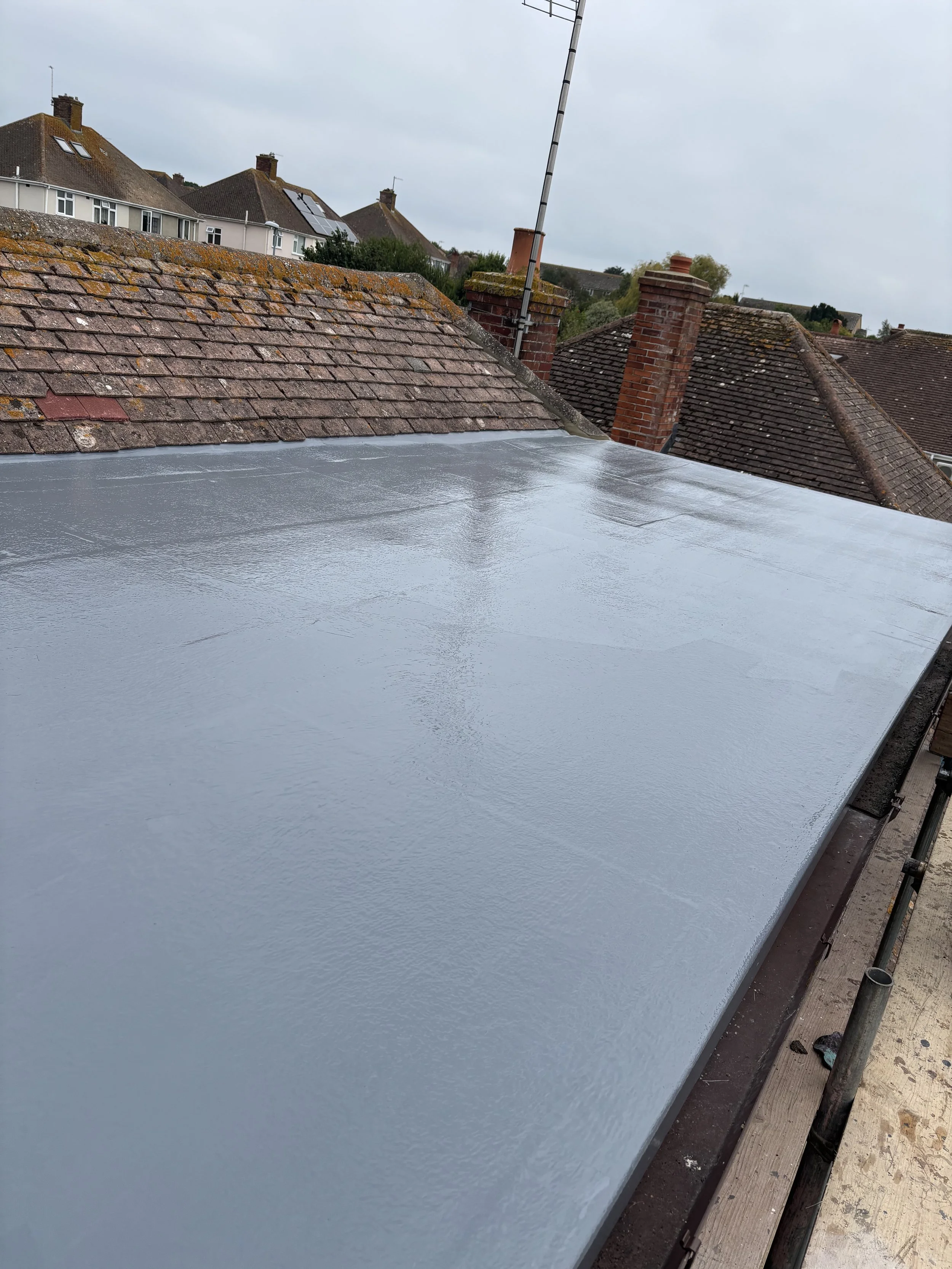 Flat roof being coated with a gray waterproofing membrane on a residential building, with neighboring rooftops and chimneys visible in a cloudy sky.