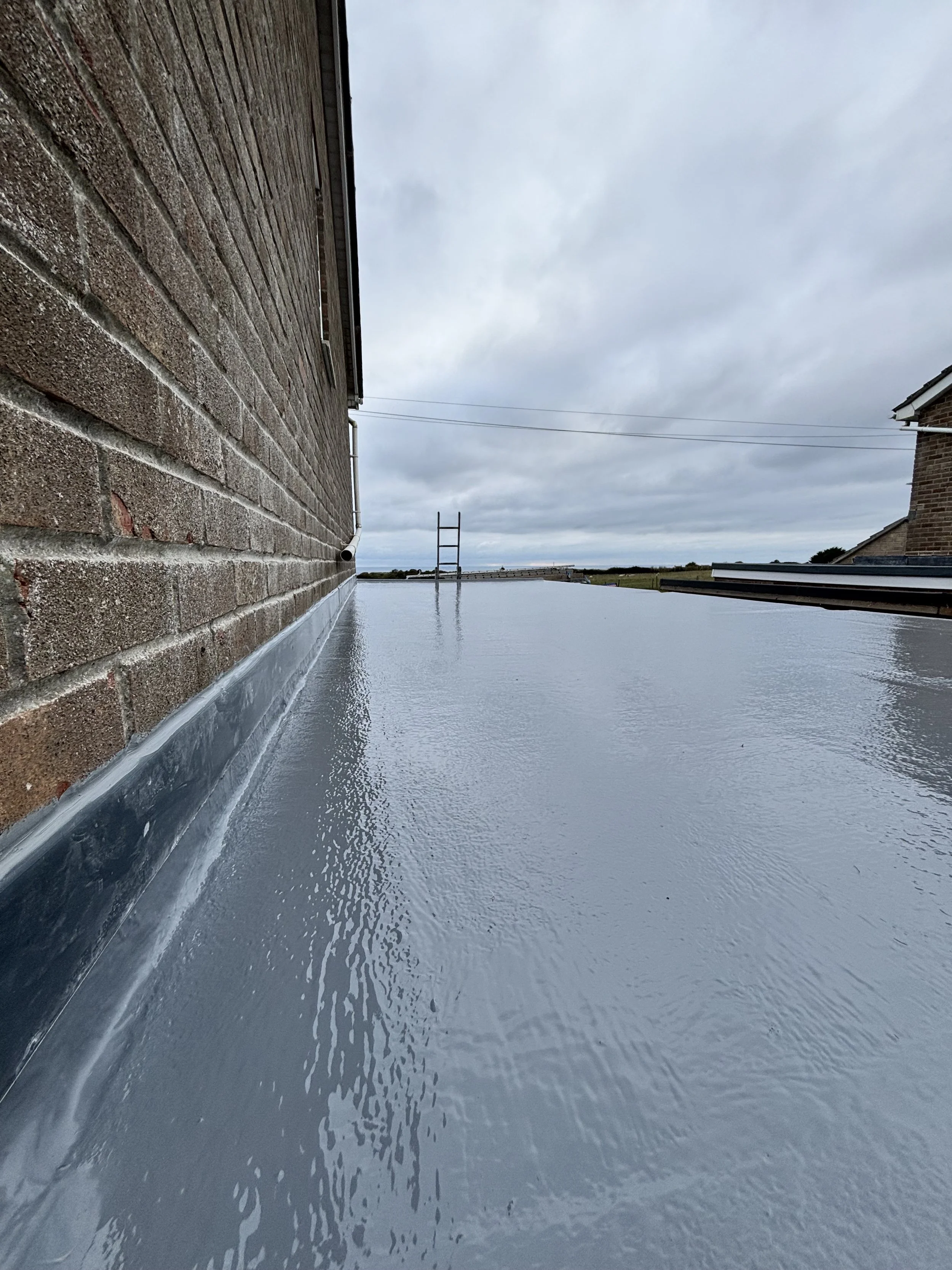 View of a flat roof with a gray waterproof coating, side of a brick house, chimney, cloudy sky, and distant power lines.