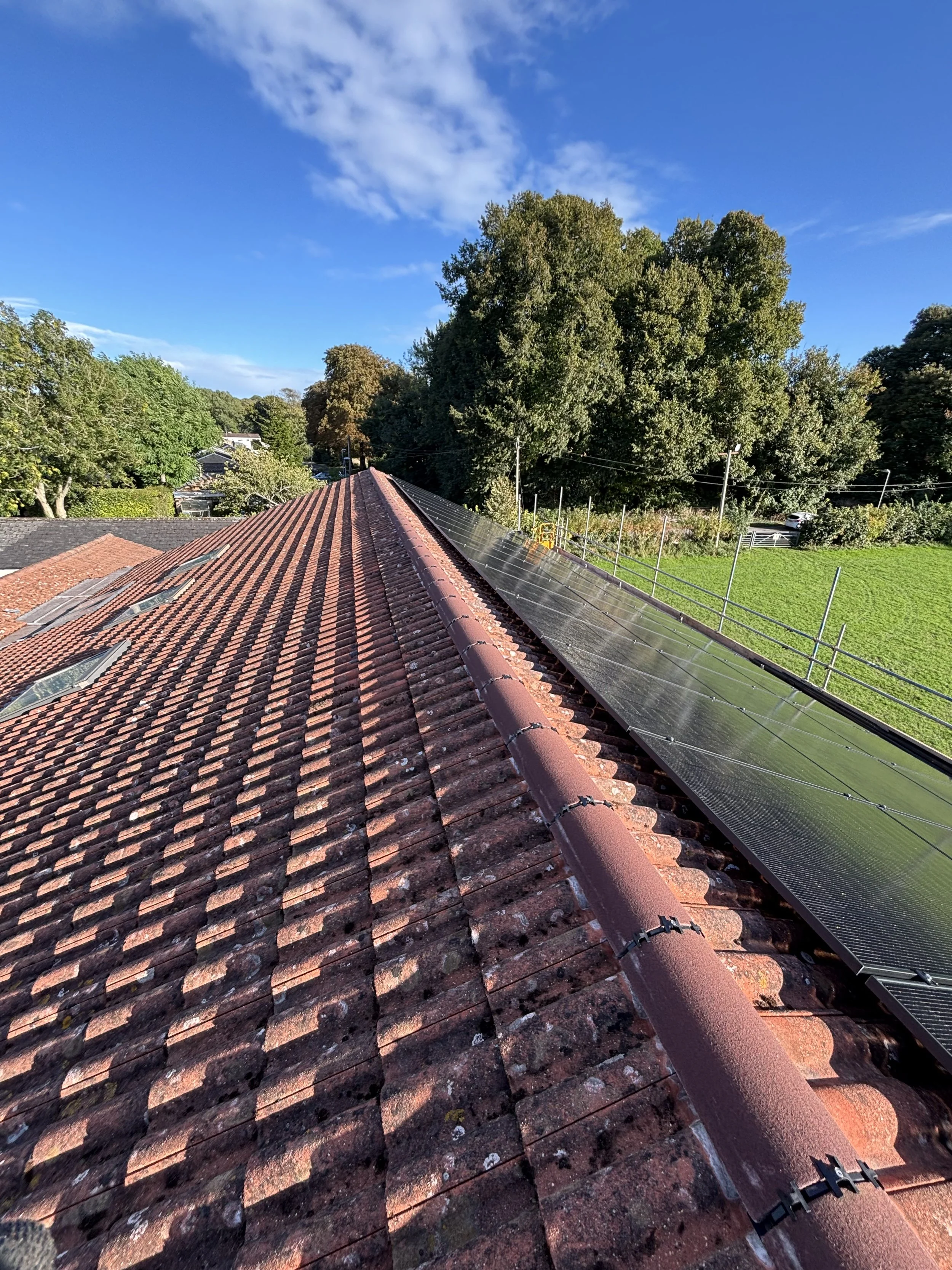 View of a rooftop with red tiles and solar panels installed along one side, with trees, a green field, and a blue sky in the background.