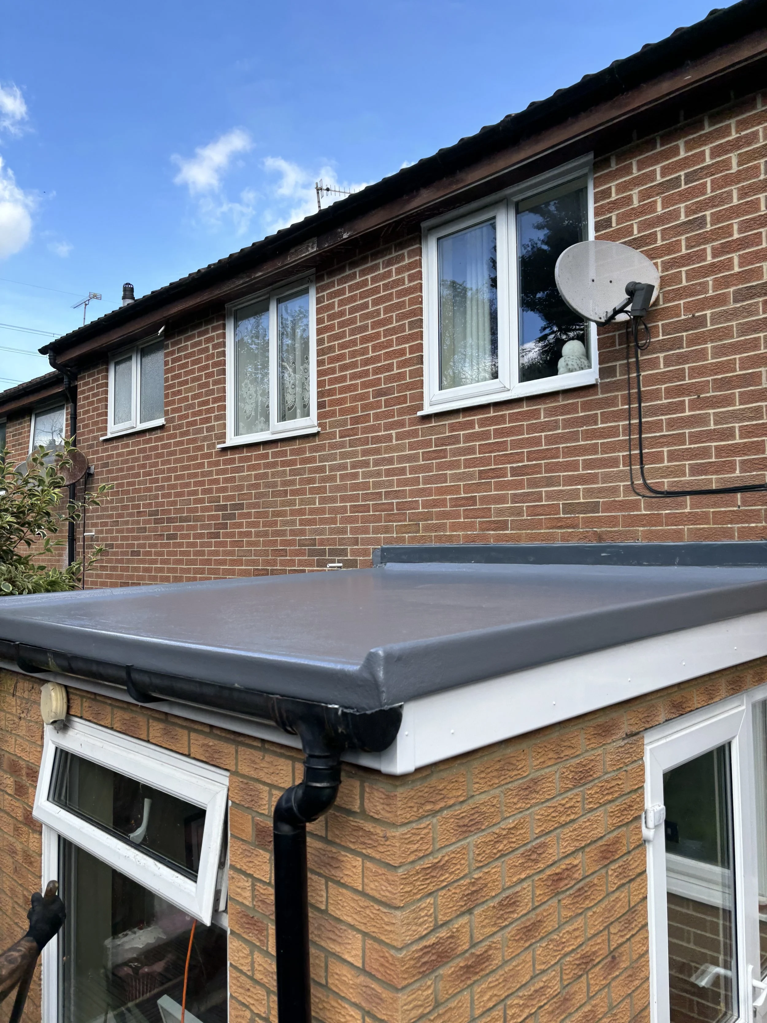 The photo shows the exterior of a two-story brick house with three windows on the upper floor, a satellite dish attached to the wall, and a small extension with a flat roof and sliding glass doors on the lower level. A person is pointing towards the window on the lower level.