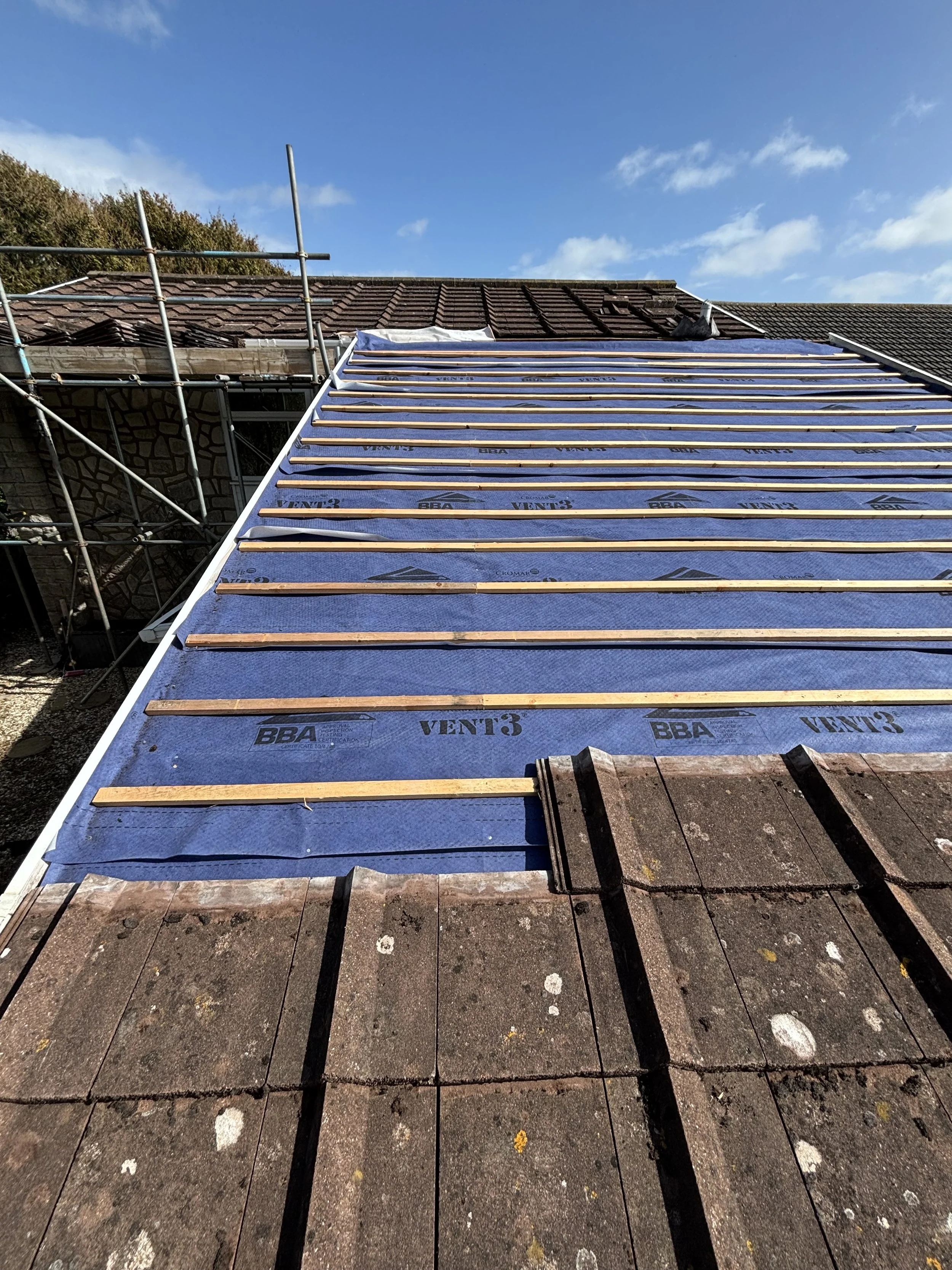 Roof under renovation showing battens and roofing underlayment, with partially removed old shingles and scaffolding on the sides under a blue sky.
