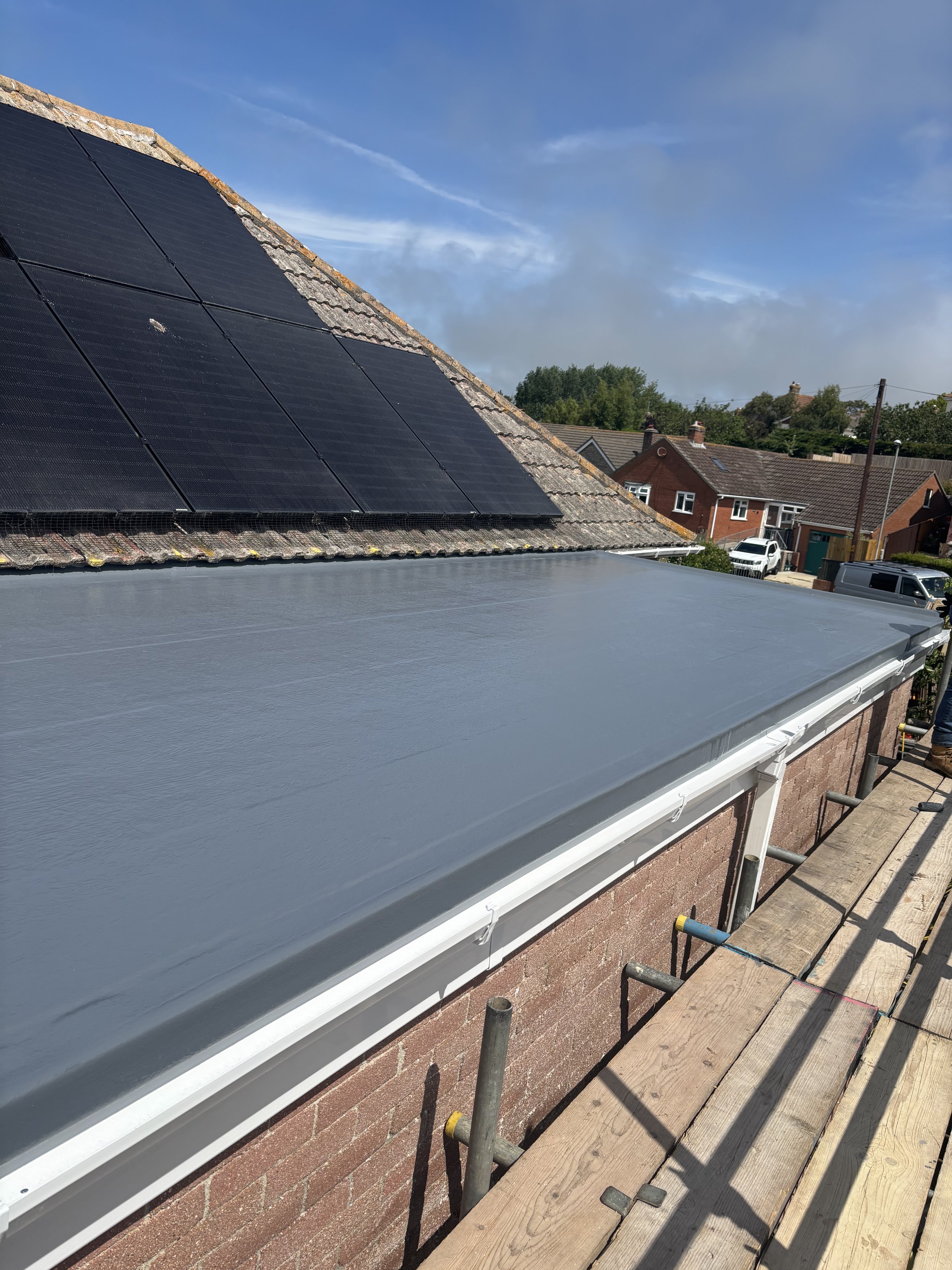 Installing new flat roofing material on a residential building with solar panels on the roof, construction scaffolding and wooden planks in the foreground, houses and trees in the background under a partly cloudy sky.