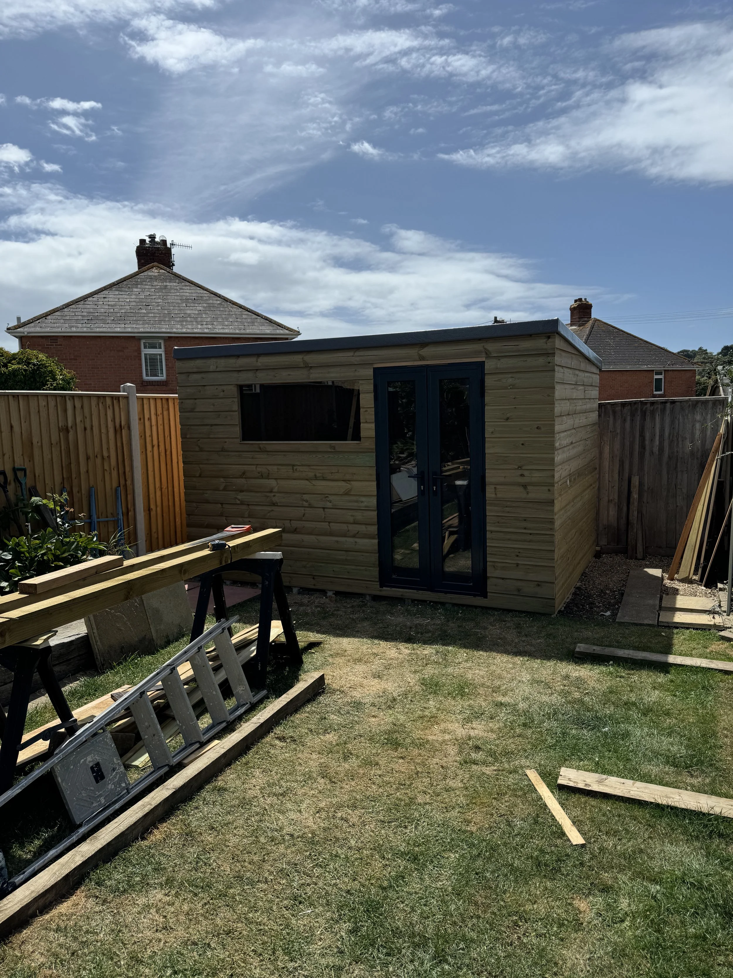 A backyard scene with a small wooden shed featuring double glass doors, surrounded by construction materials, with two neighboring brick homes and a blue sky with clouds in the background.