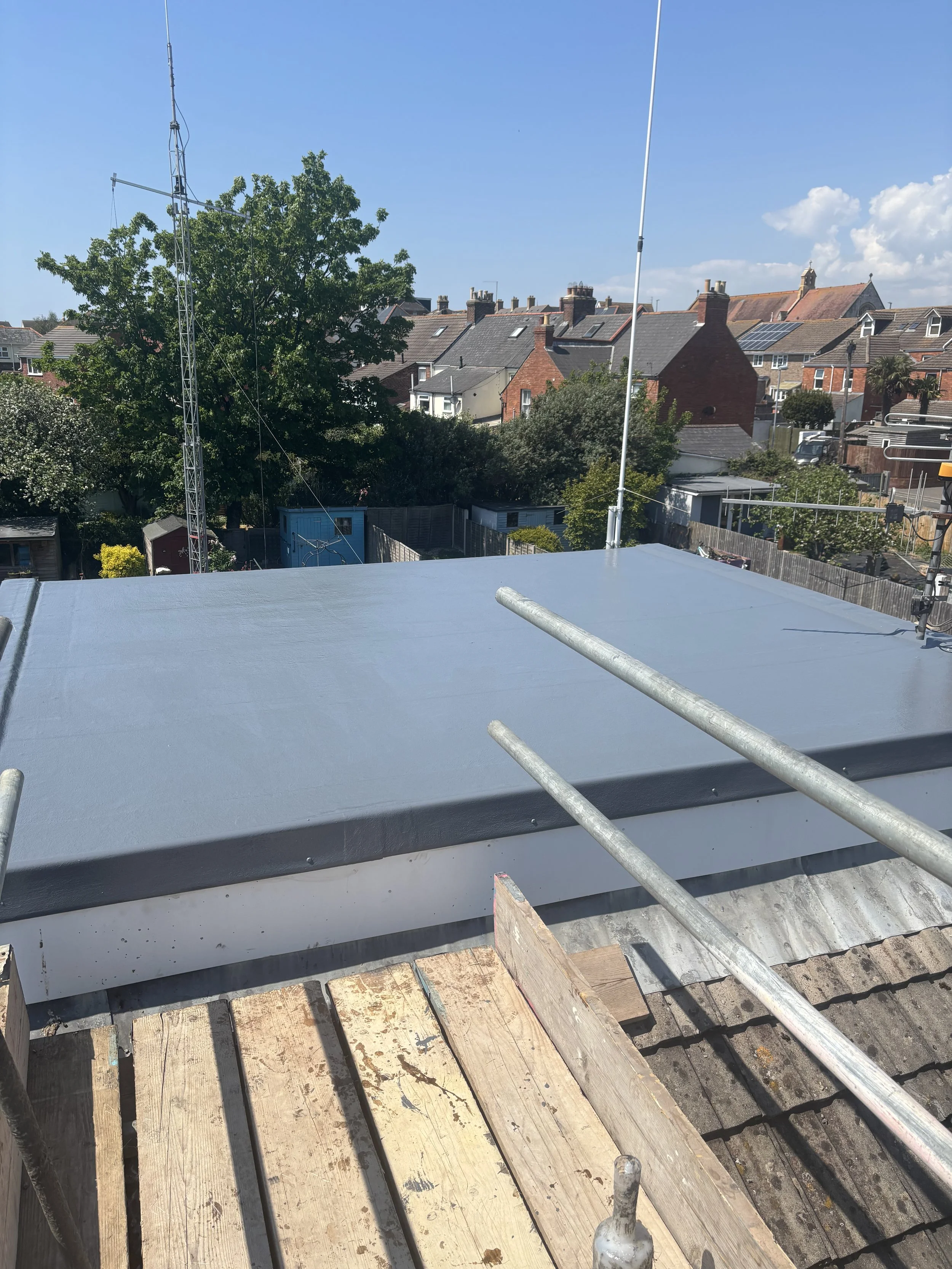 Roof undergoing repairs with a fresh coat of gray paint, wooden planks, and scaffolding. Residential houses and trees in the background under a clear blue sky.
