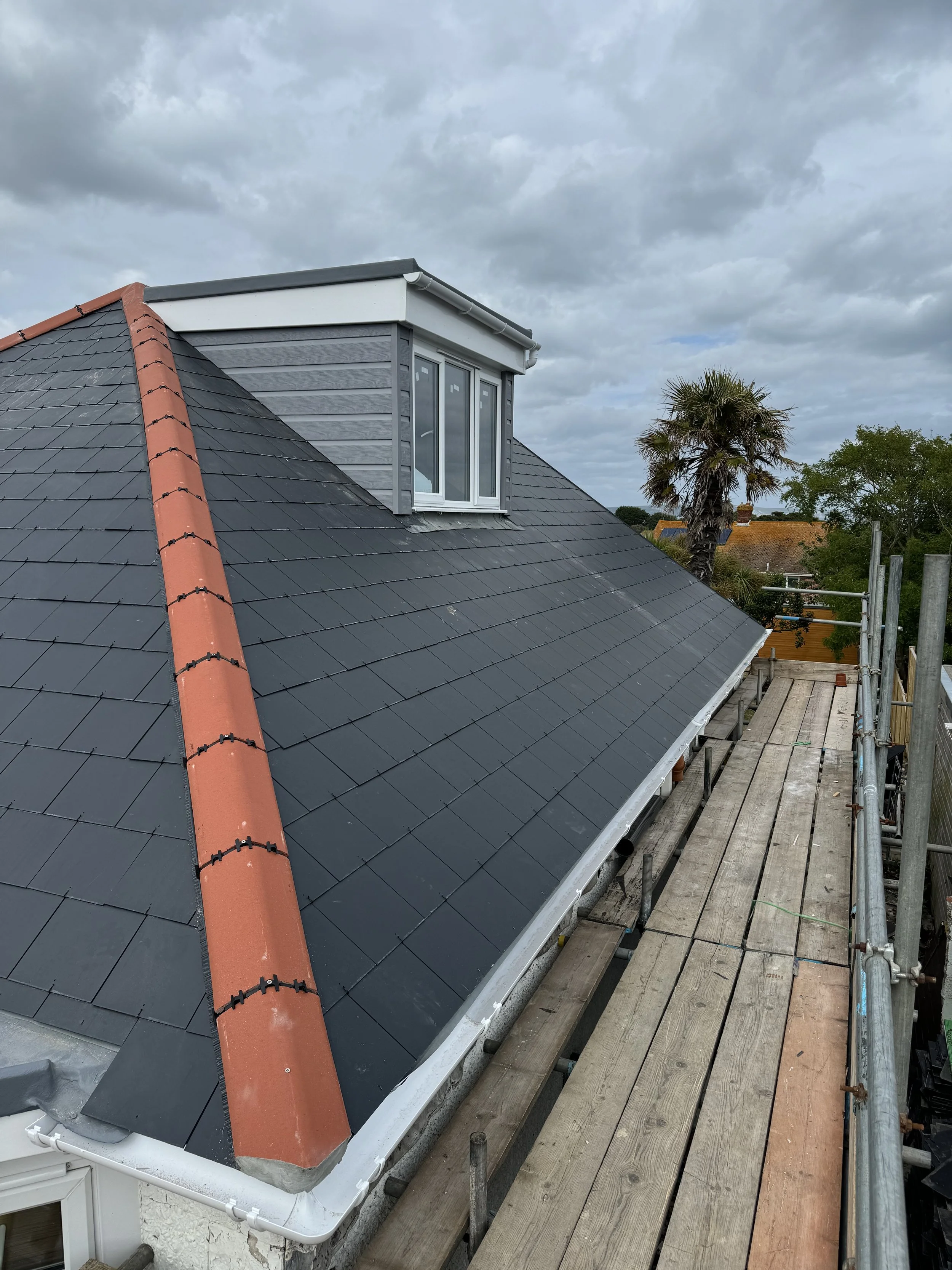 View of a dark gray slate roof with a red ridge tile, a dormer window protruding from the roof, and cloudy sky in the background. Construction scaffolding is along the edge of the roof.