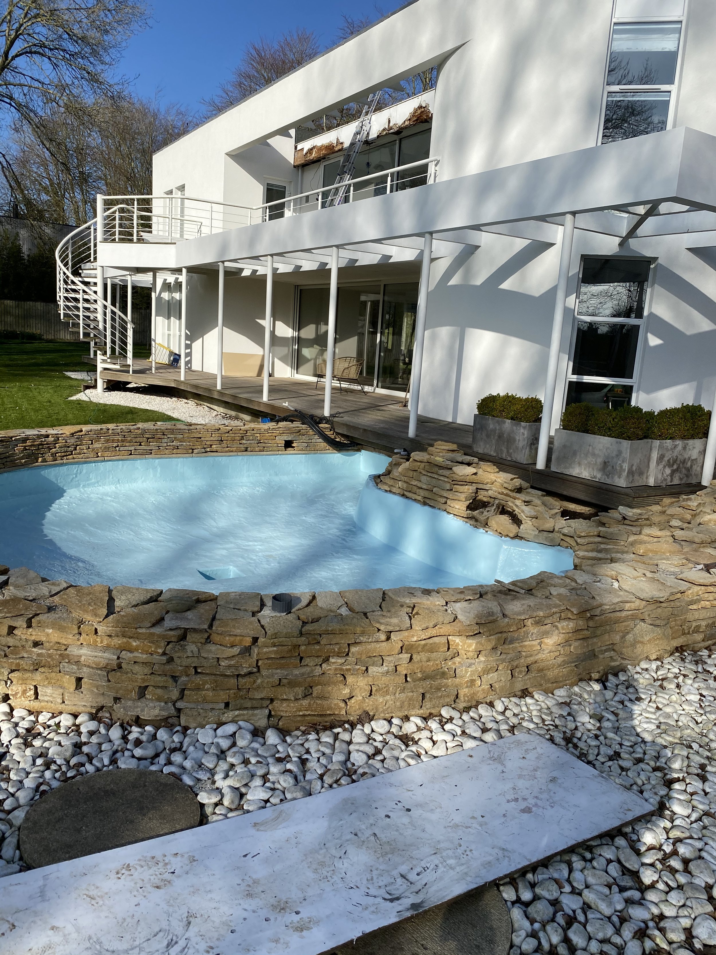 Modern white house with a balcony, a spiral staircase, a deck area, and a small in-ground pool surrounded by stone and gravel landscaping.