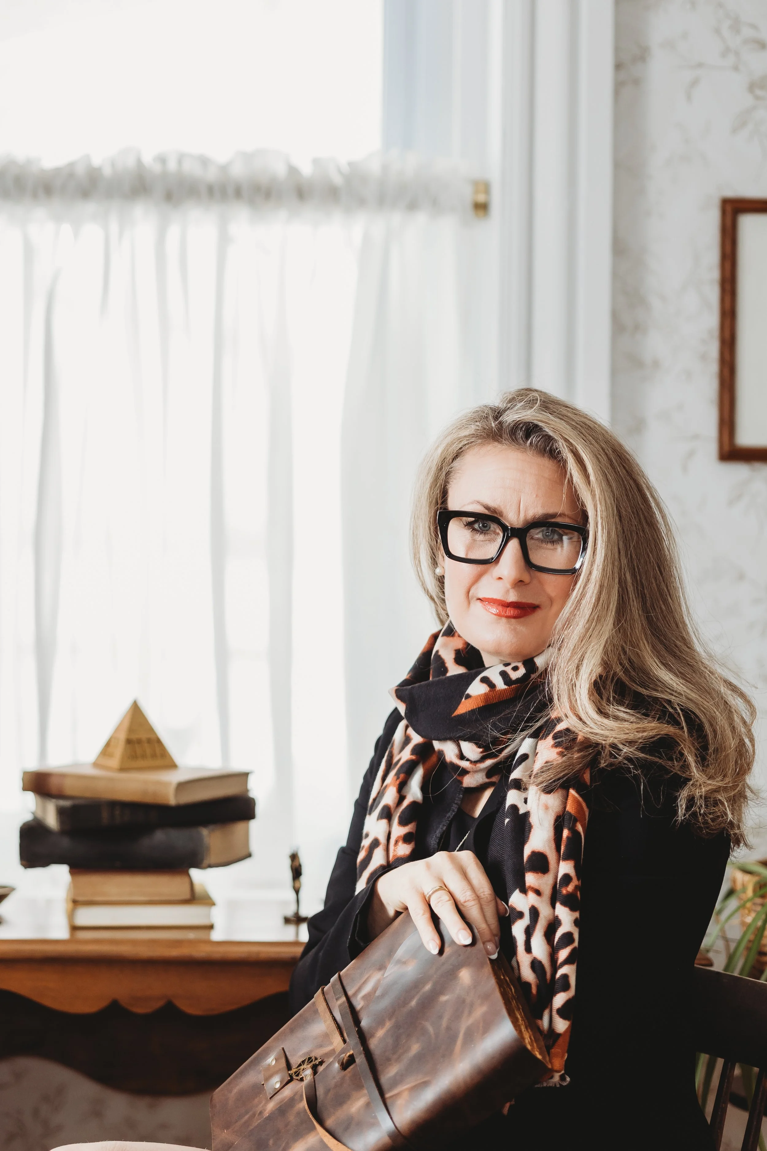 A woman with blonde hair, glasses, and a scarf, sitting in a well-lit room with books and decor, holding a bag and looking at the camera.
