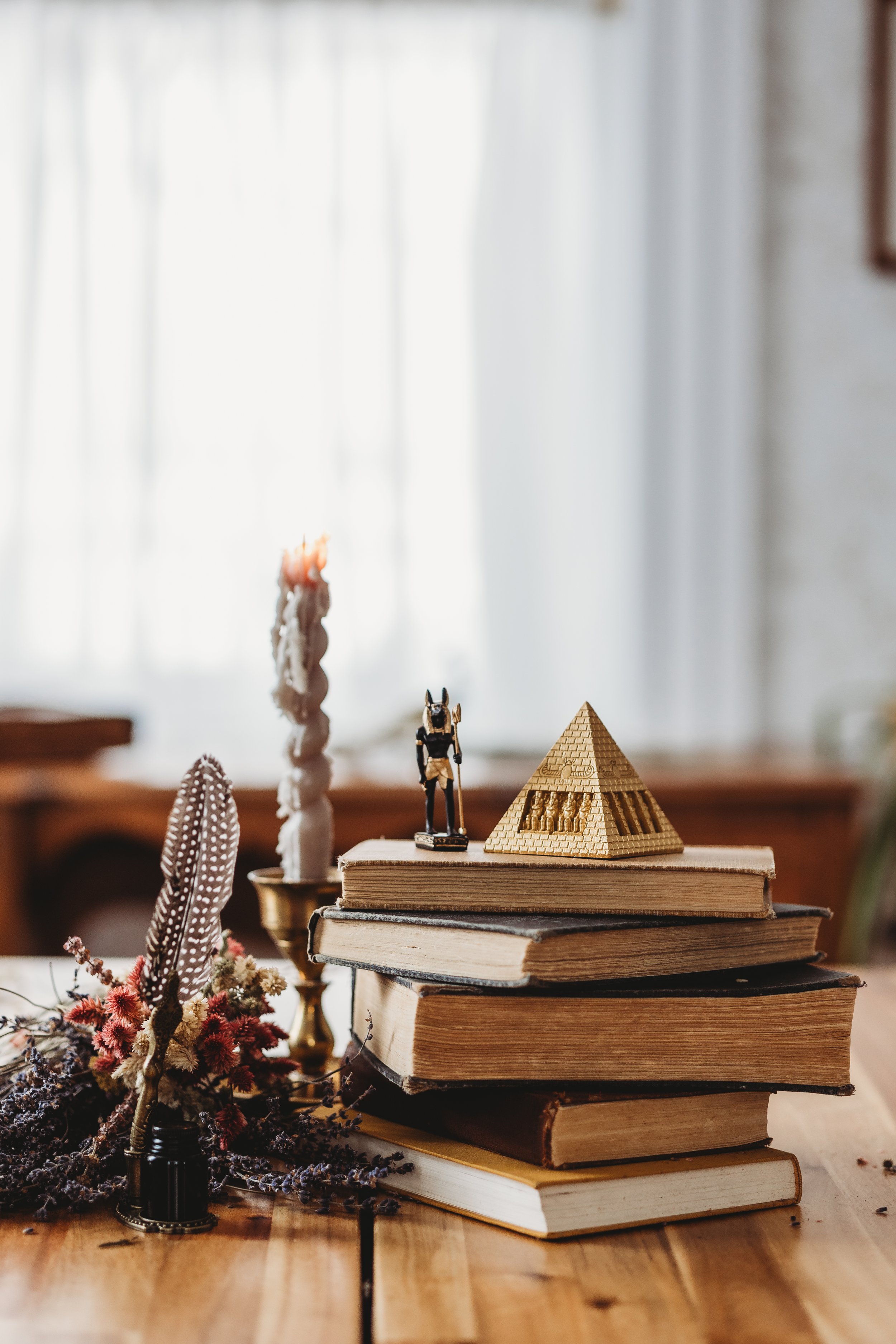 Stack of old books on a wooden table with decorative items including a feather, flowers, a small figurine, and a pyramid-shaped object.