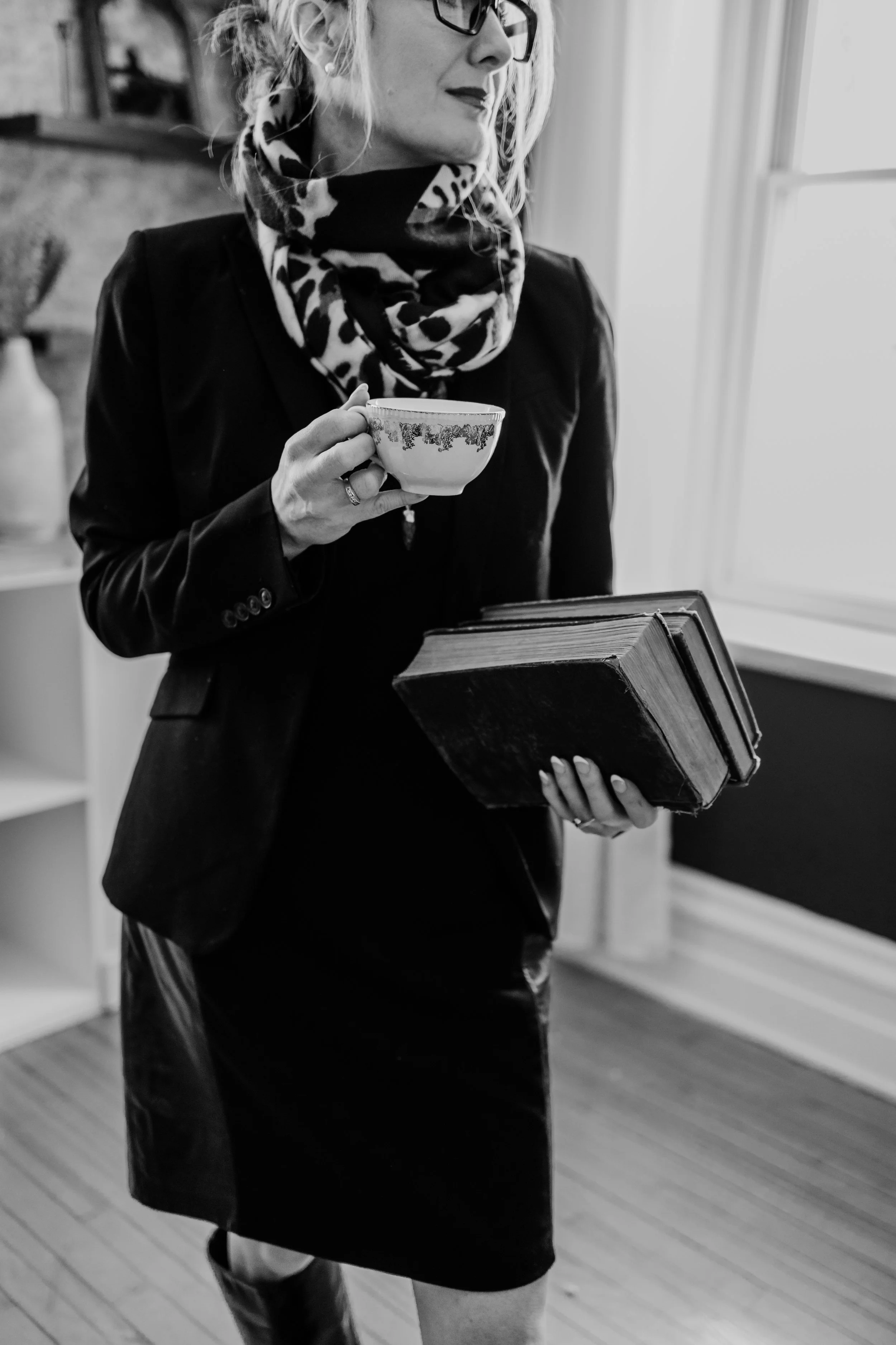 A woman wearing glasses, a leopard-print scarf, a black blazer, and a black skirt, holding a teacup in one hand and a stack of three books in the other, standing by a window in a room with wooden floors and shelves.