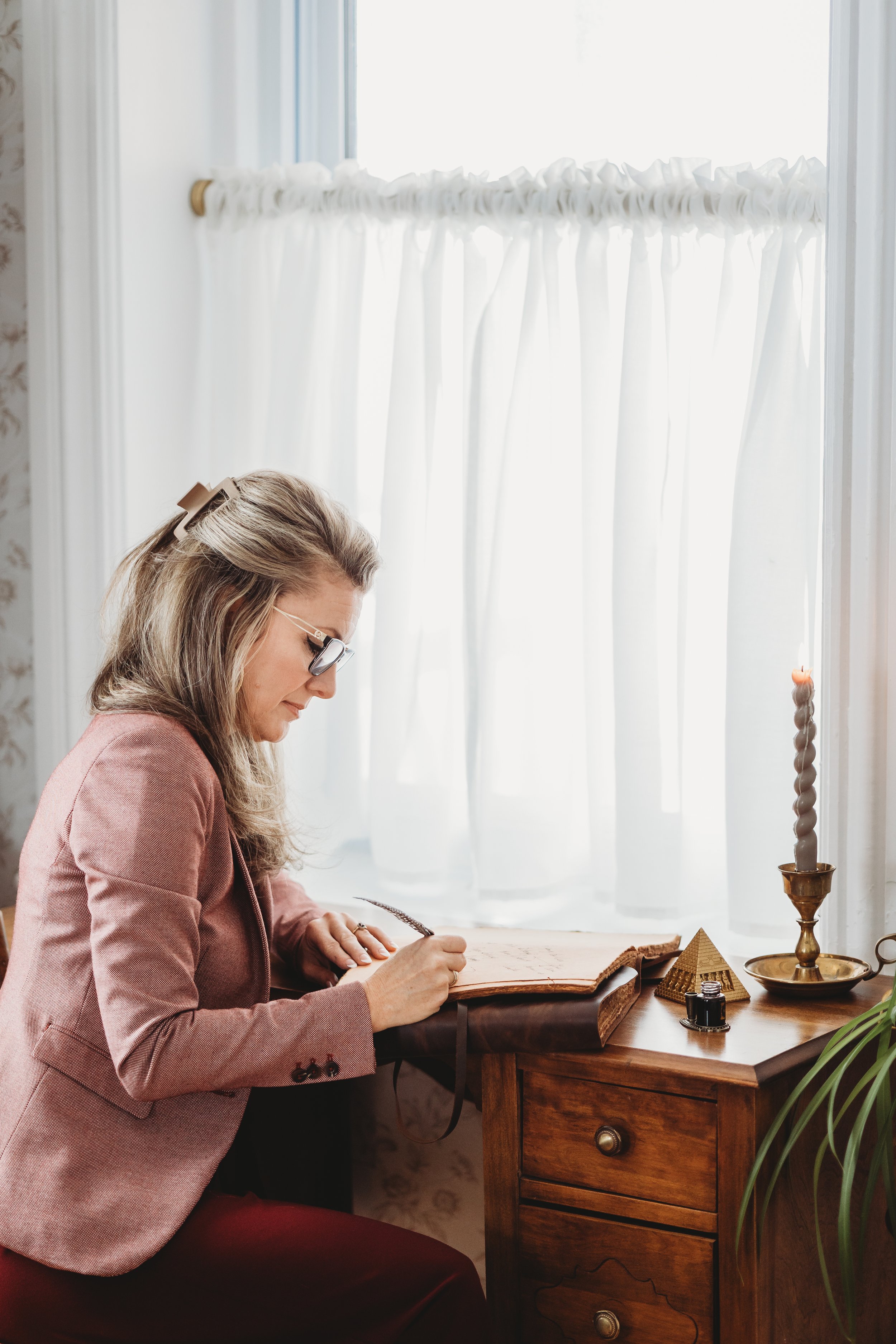 Woman with glasses writing in a large notebook at a wooden desk, near a window with white curtains and a candle holder with a lit candle.