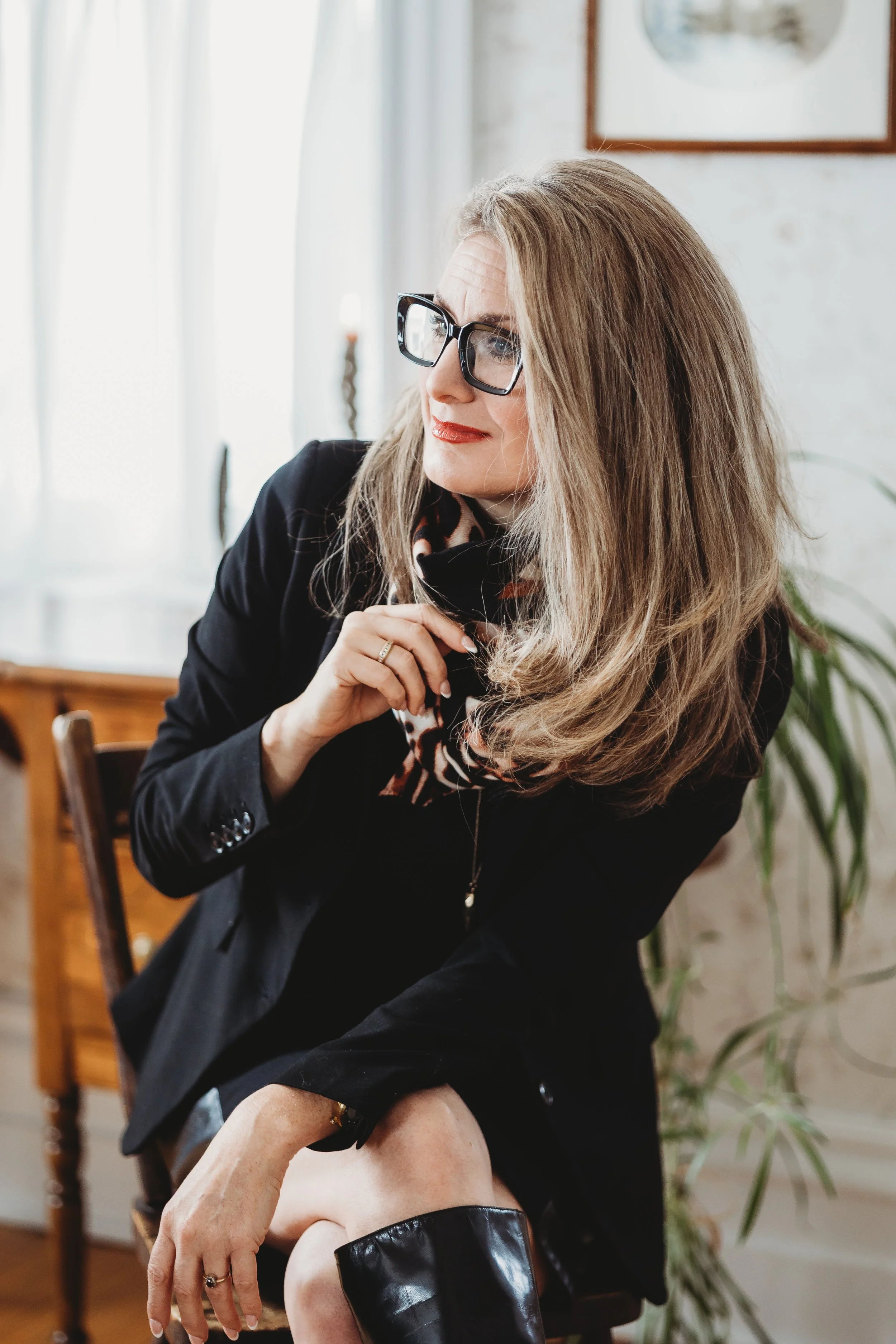 A woman with long, wavy blonde hair wearing black glasses, a black blazer, a patterned scarf, and knee-high black boots, sitting on a wooden chair in a well-lit room with a plant and framed pictures in the background.