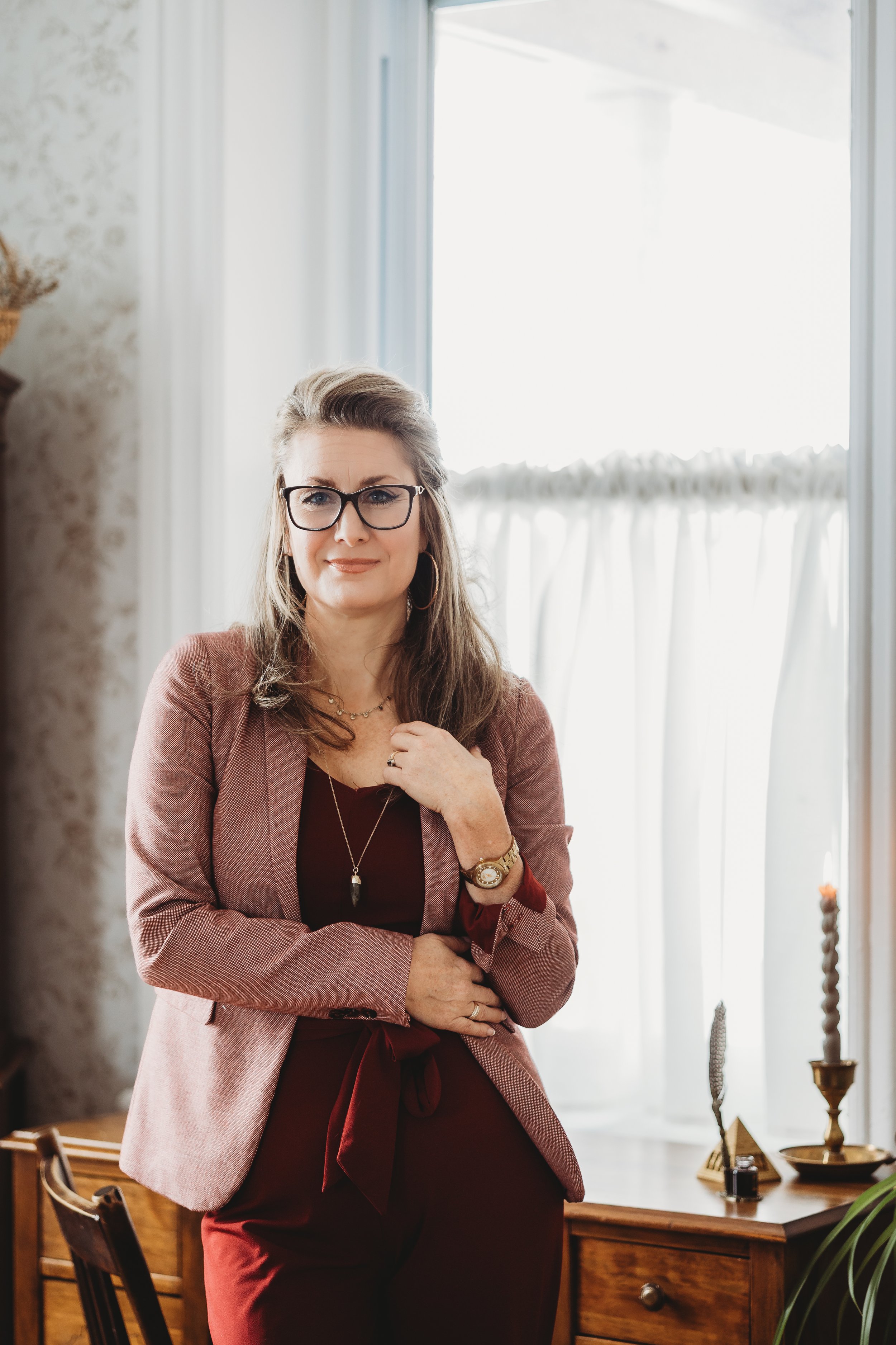 A woman standing indoors near a window with sheer curtains, wearing glasses, a pink blazer, and a maroon dress, with jewelry and a watch, looking at the camera.