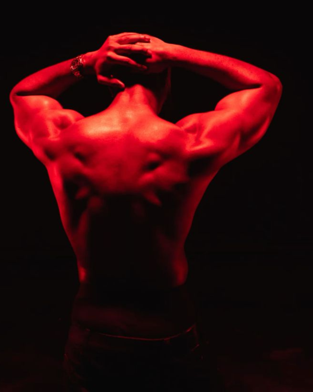Back view of a muscular person with red lighting, arms raised and hands on head, against a black background.