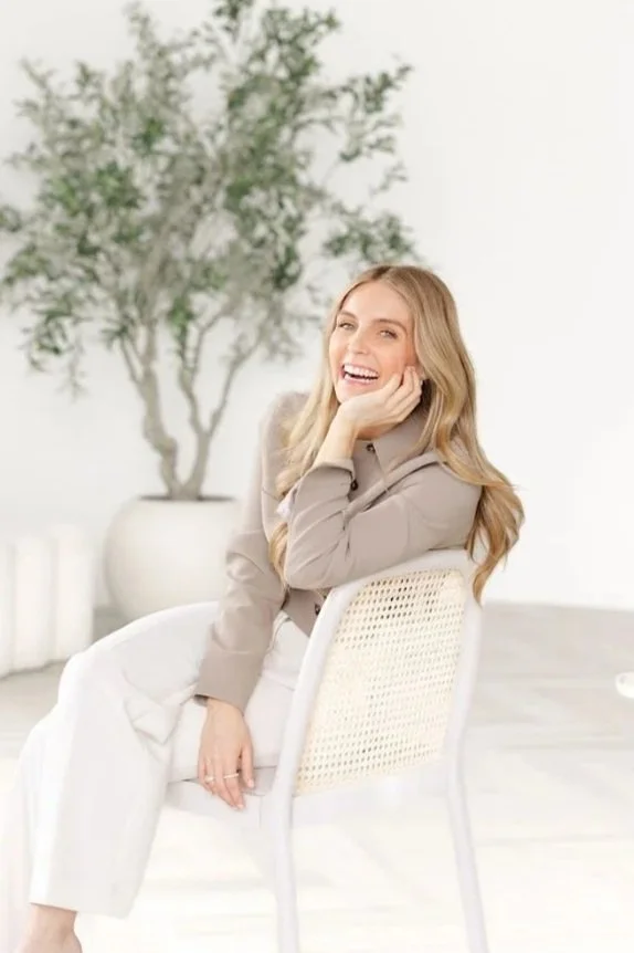 A young woman with long blonde hair sitting on a white chair, smiling and resting her chin on her hand, in a bright room with a large potted plant in the background.