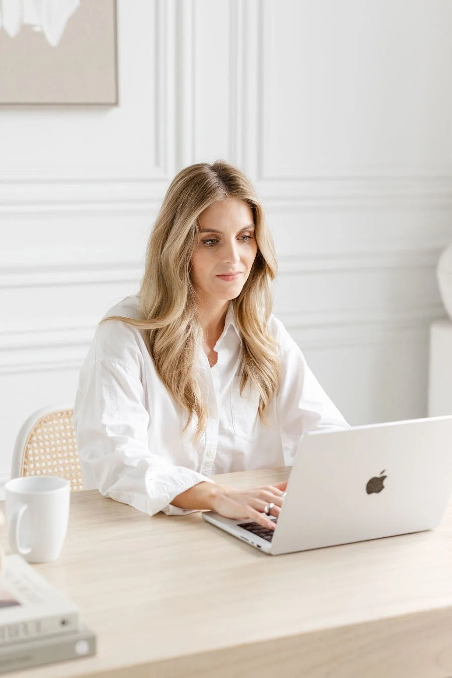 Woman working on a MacBook at a light wood desk in a bright, white room.