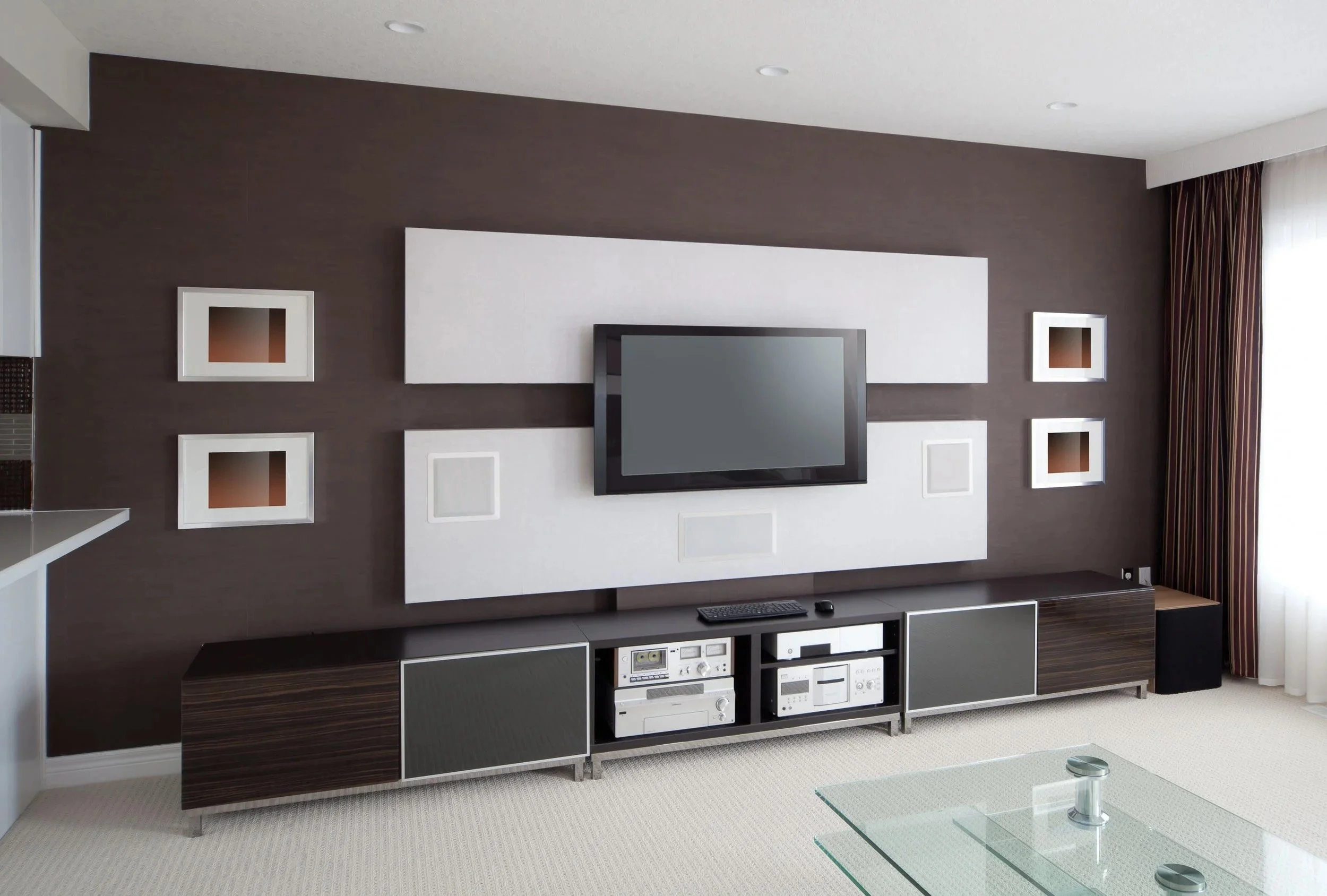 Modern living room with a wall-mounted flat-screen TV on a white wall panel, surrounded by decorative square frames, dark brown wall, beige carpet, and glass coffee table.