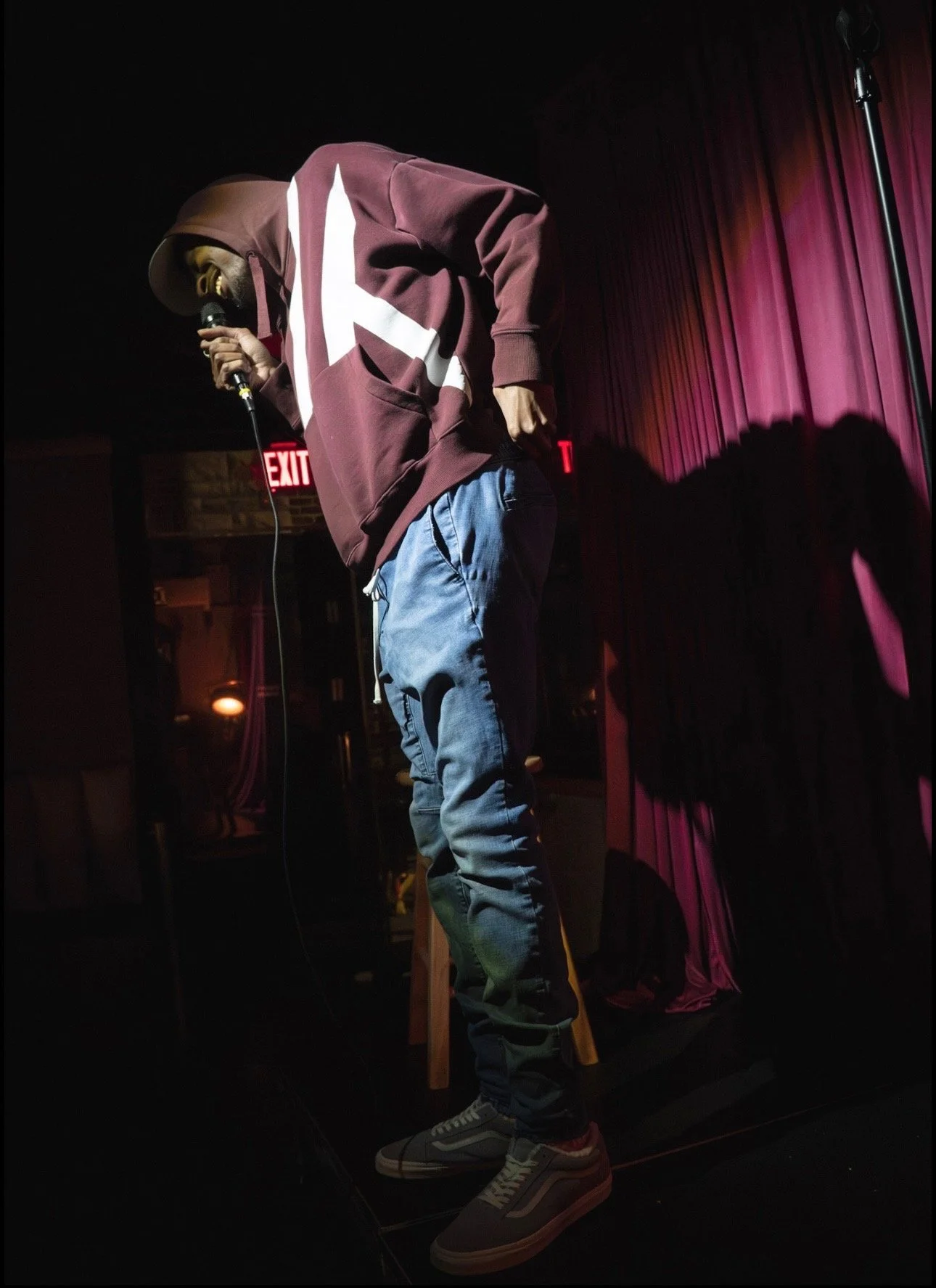RJ performing on stage with a microphone, wearing a hoodie, jeans, and sneakers, against pink curtains and a black background.
