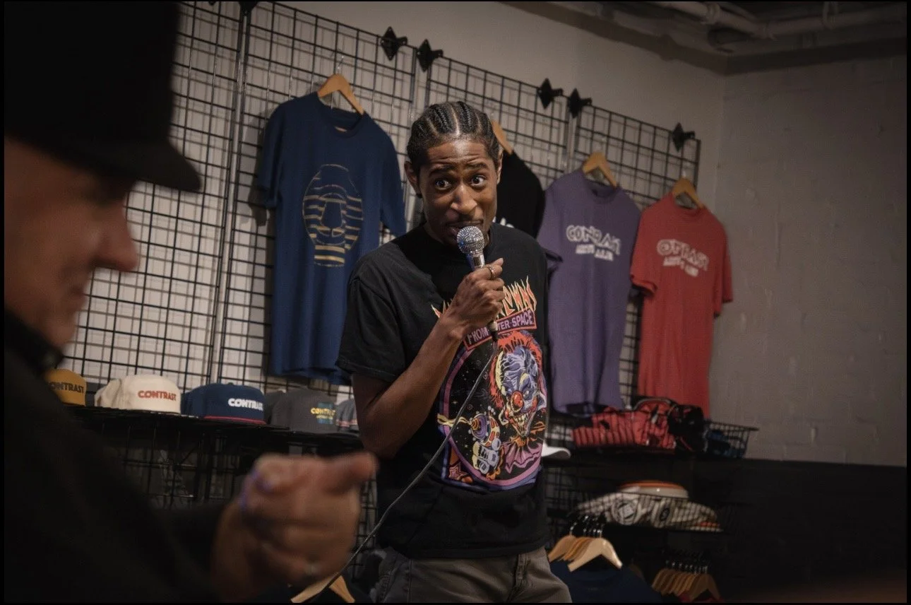 RJ holding a microphone, speaking to an audience in a retail store with shirts and caps on display in the background.