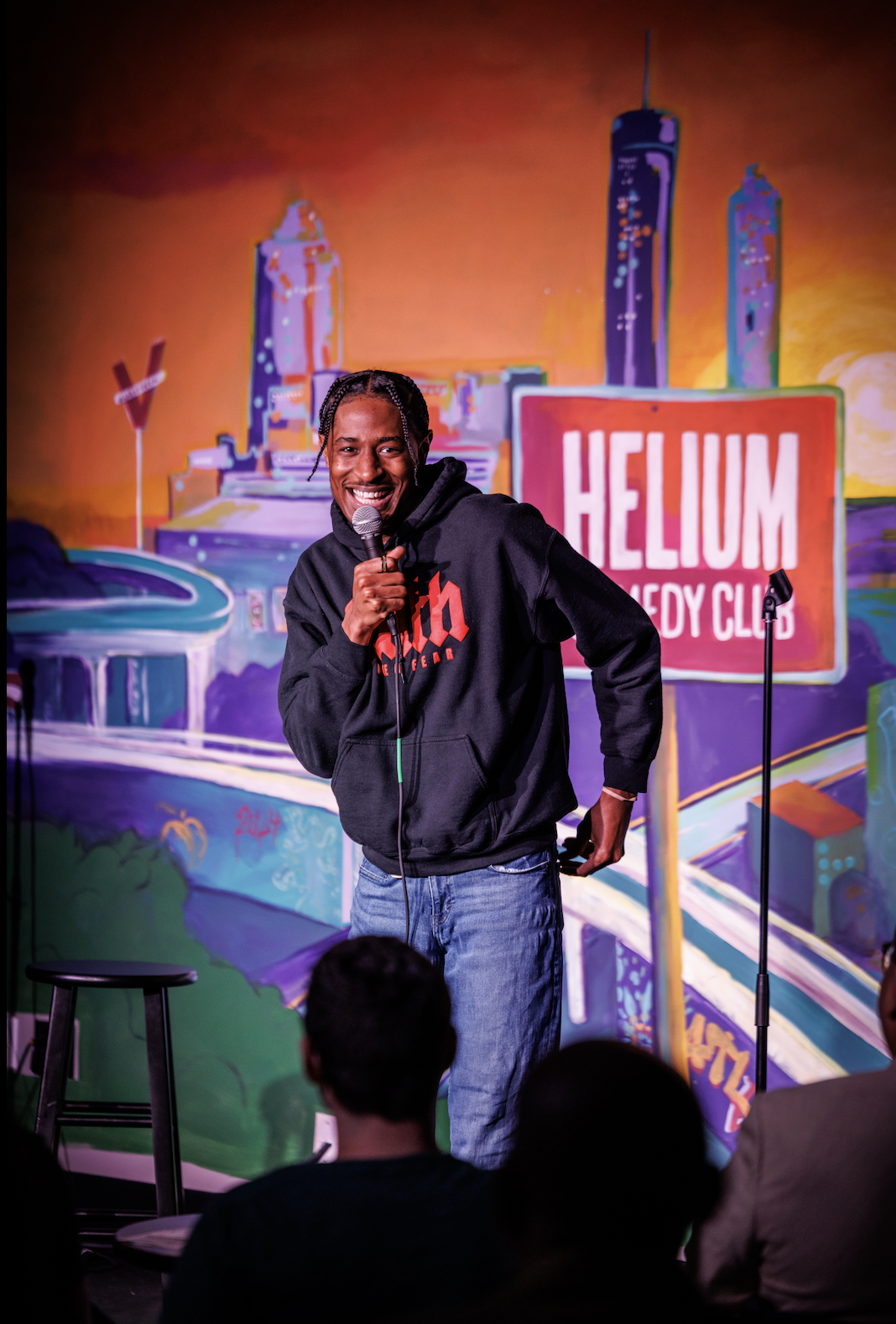 RJ performing stand-up comedy on stage at Helium Comedy Club, holding a microphone and smiling, with a colorful cityscape mural background and audience members in the foreground.