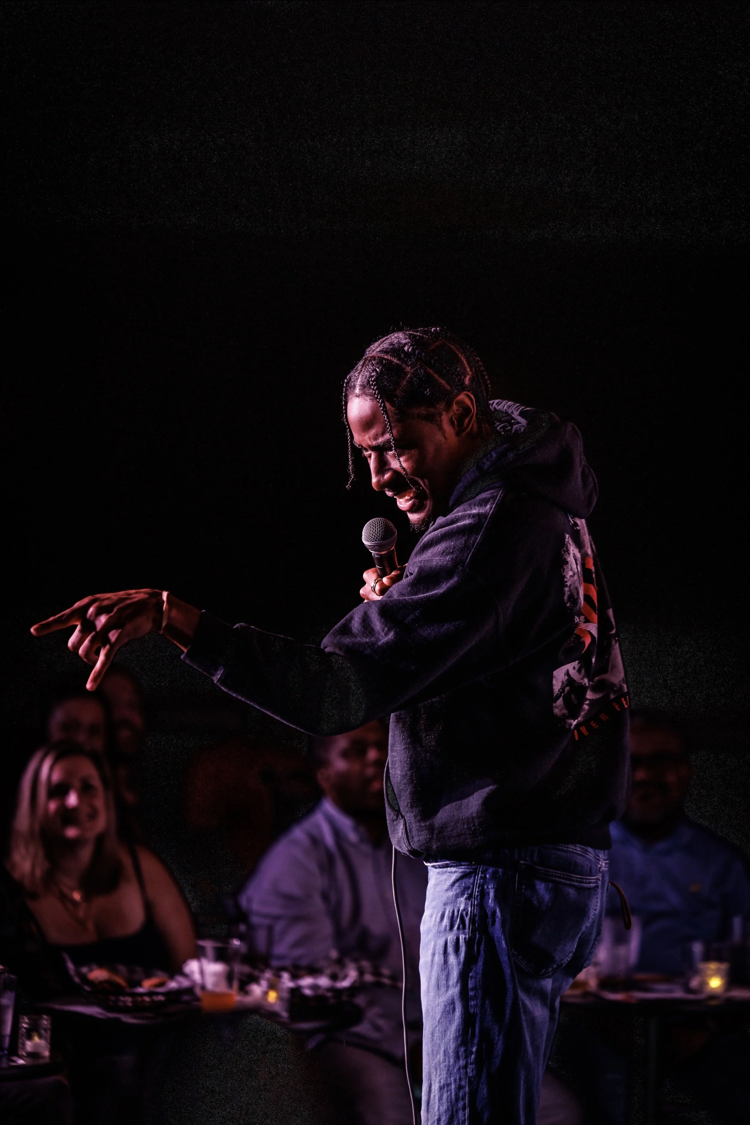 RJ the Character with braided hair performing stand-up comedy with a microphone, crowd watching and reacting at a comedy venue.