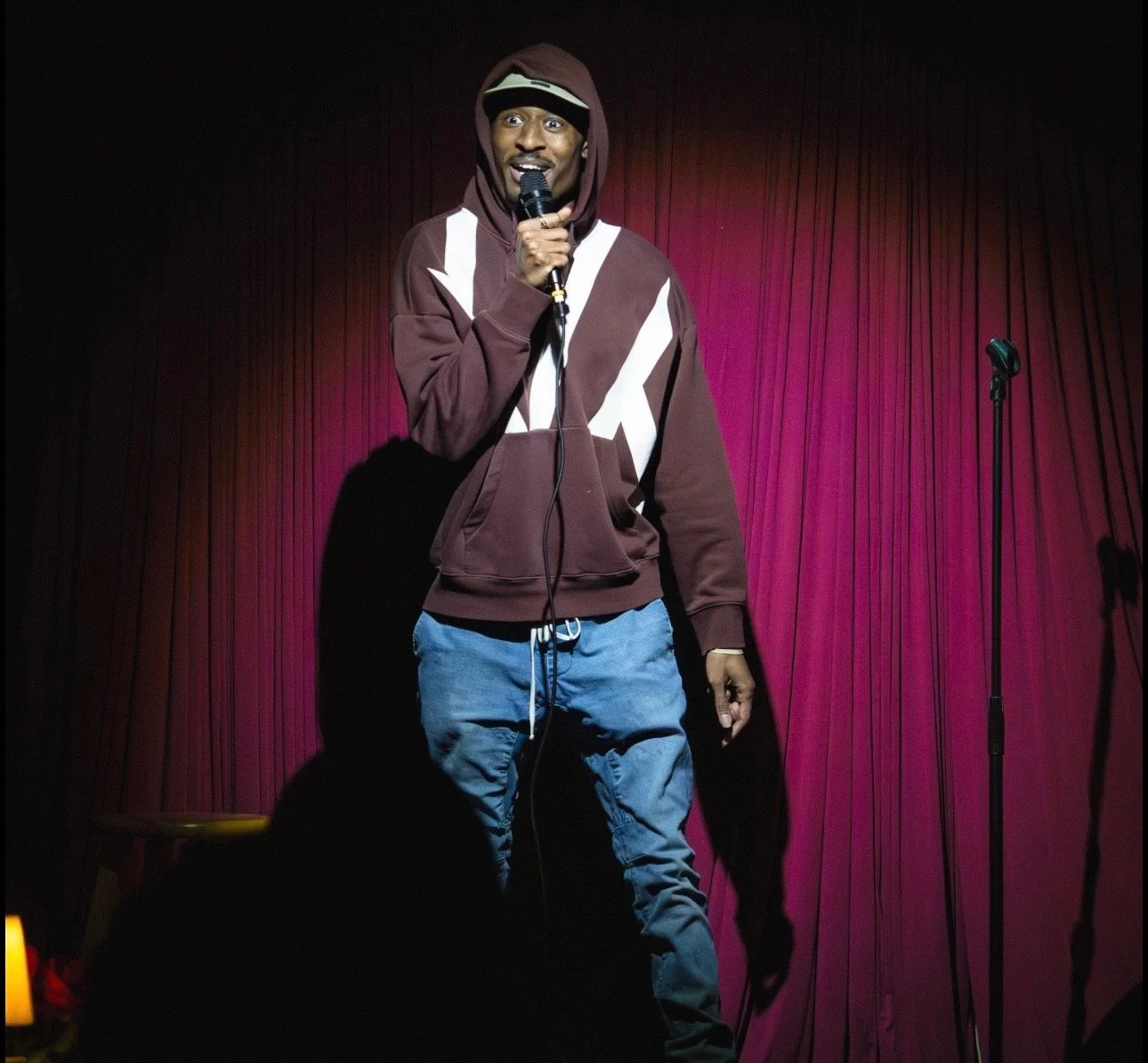RJ performing stand-up comedy on stage with a microphone in front of a pink stage curtain, wearing a hoodie and jeans.