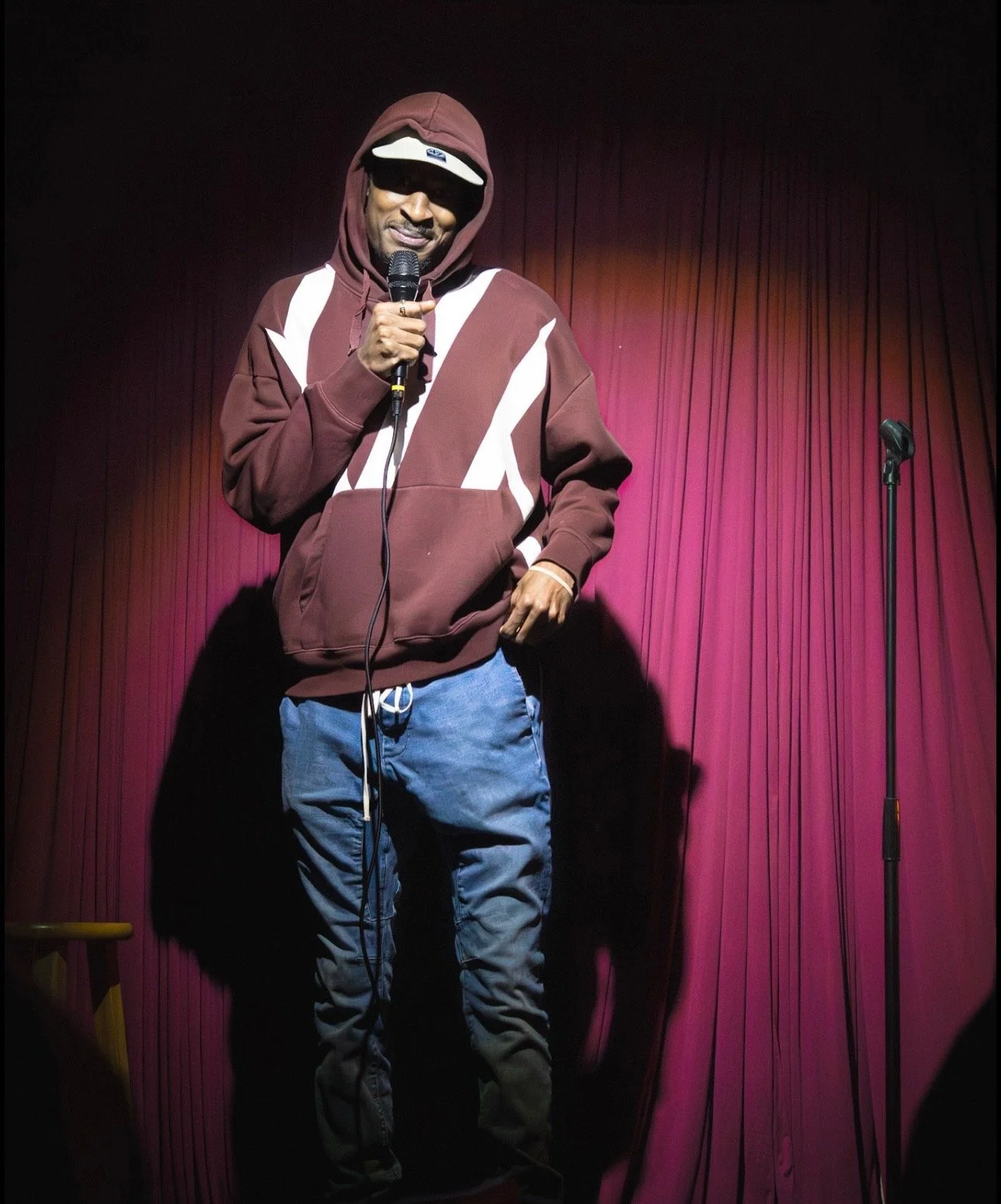 RJ wearing a brown hoodie, sunglasses, and a beige cap speaks into a microphone on a stage with pink curtains.