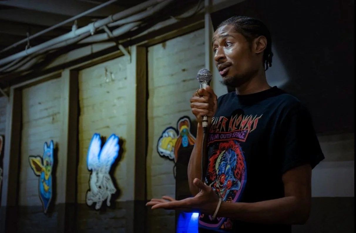 RJ the Character, holding a microphone and speaking in front of a mural wall with cartoon characters.