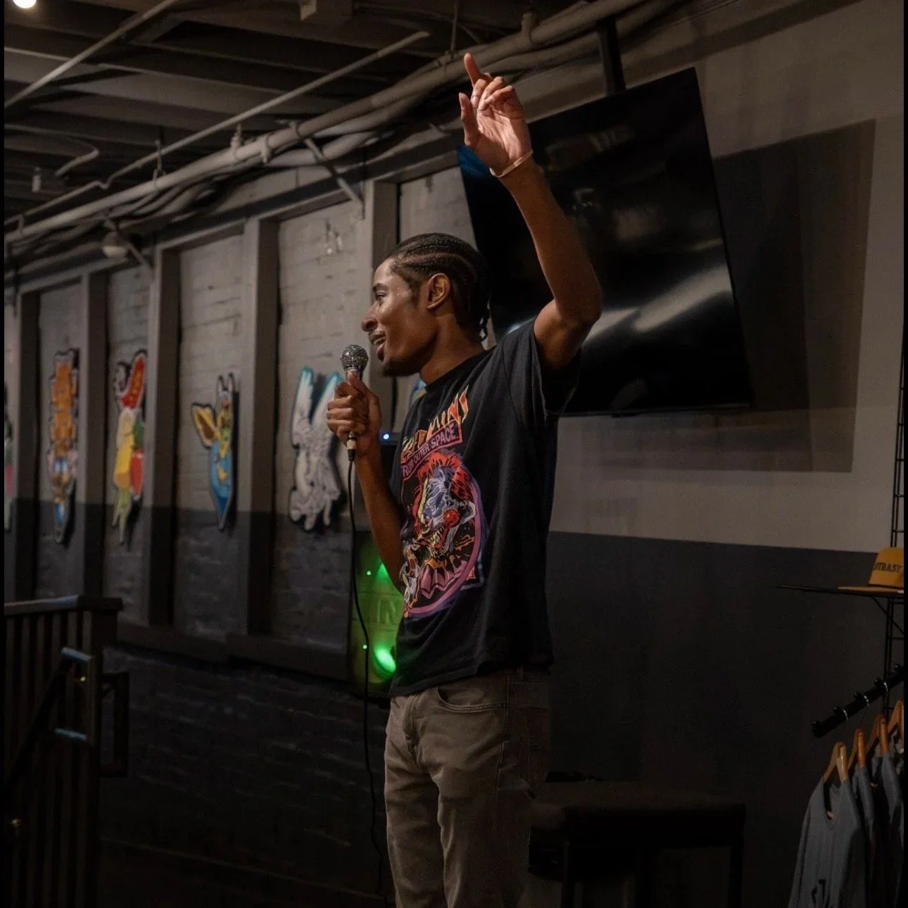 RJ the Character, wearing a black graphic T-shirt and beige pants, is holding a microphone and speaking in a dimly lit room with black and white walls. RJ is raising one hand.
