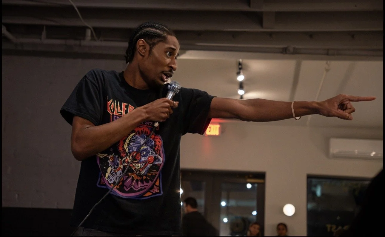 RJ the Character, holding a microphone and pointing to his right in a room with a gray ceiling and visible light fixtures.