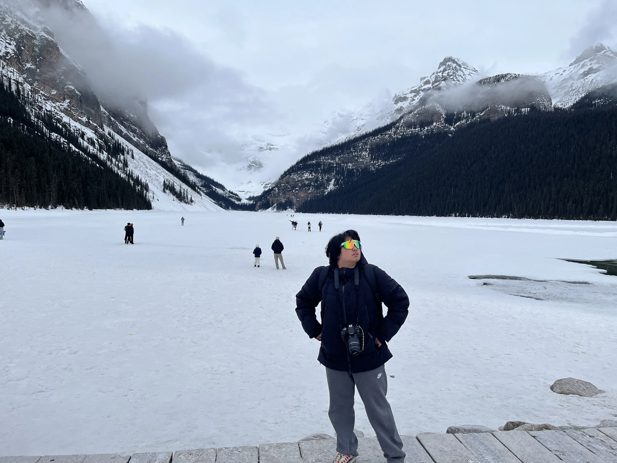 Person in black jacket and gray sweatpants with camera hanging around neck, wearing reflective sunglasses, standing on wooden pathway near snow-covered frozen lake surrounded by snow-capped mountains and pine trees, with cloudy sky.