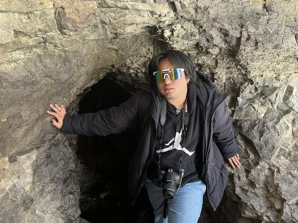 A young man wearing colorful sunglasses, a black hoodie, and a black jacket is crouching in front of a small cave or tunnel through a rocky formation. He has a camera hanging around his neck and feels like he is exploring or hiking.