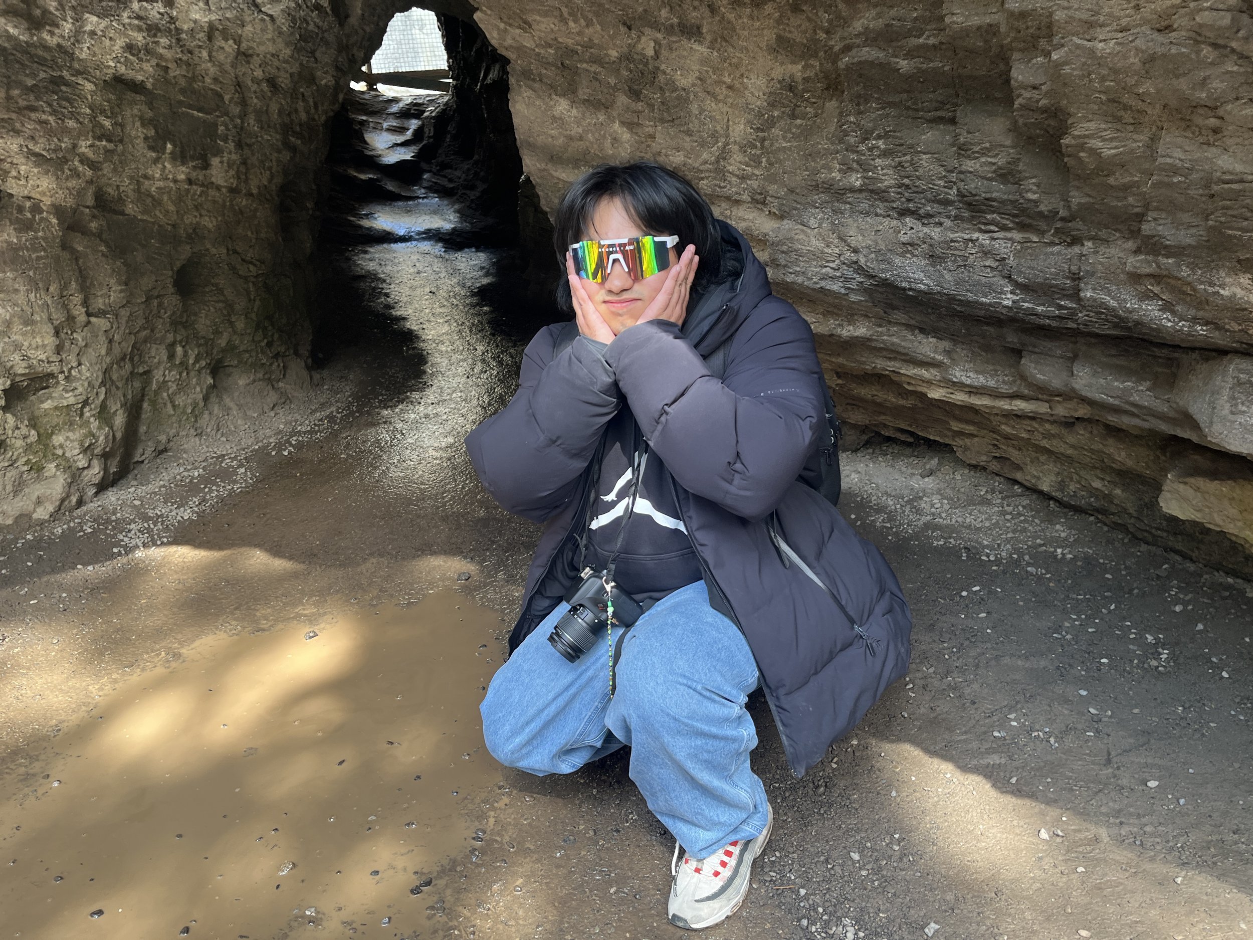 Person wearing a black jacket, gray sweatpants, colorful sunglasses, and a camera hanging around their neck crouching in front of a small cave or crevice in rocky terrain with a small puddle of water nearby.