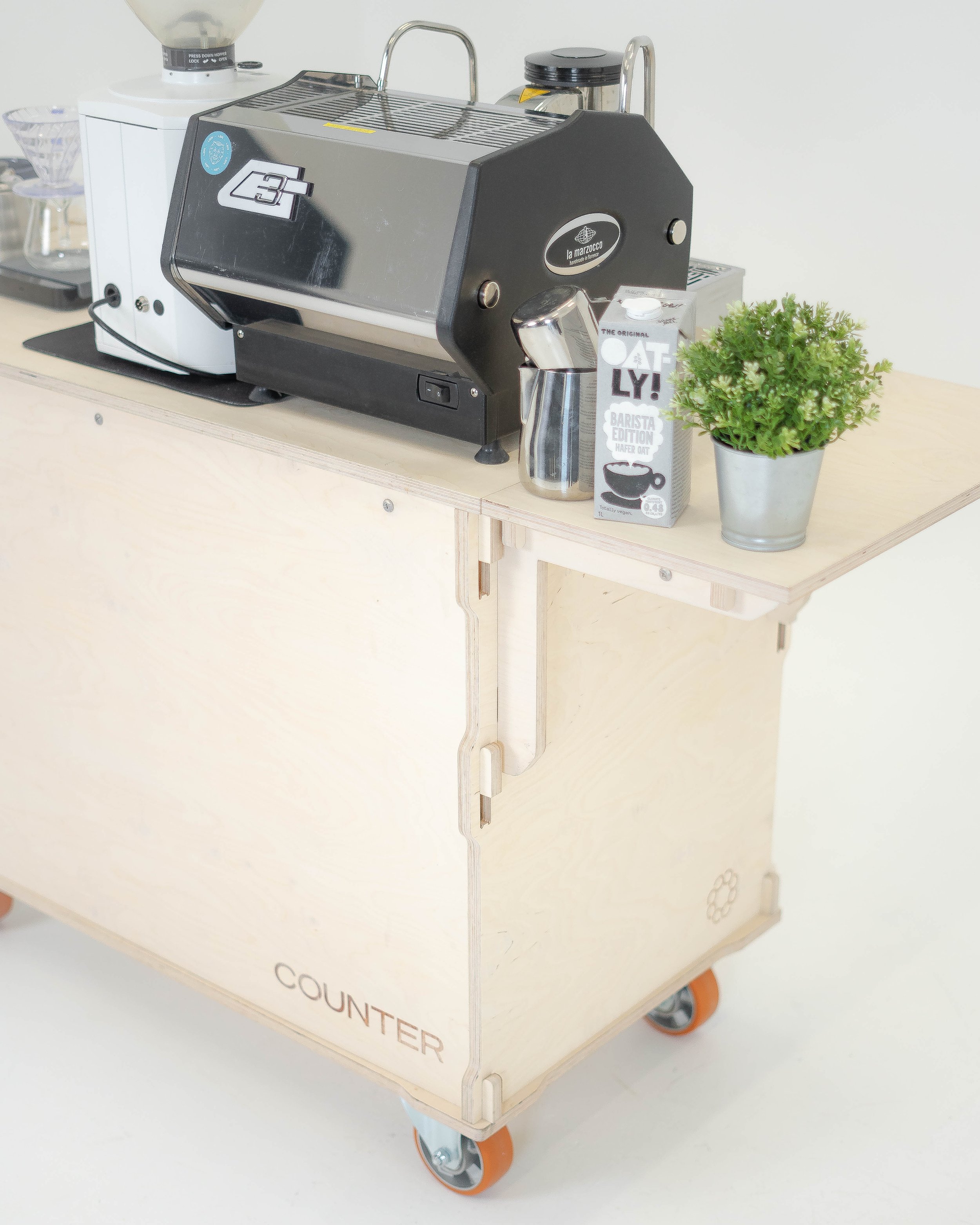 A wooden cart with the word 'COUNTER' printed on its side, equipped with a coffee roasting machine, a potted plant, a coffee scoop and a carton of oat milk, on a white background.